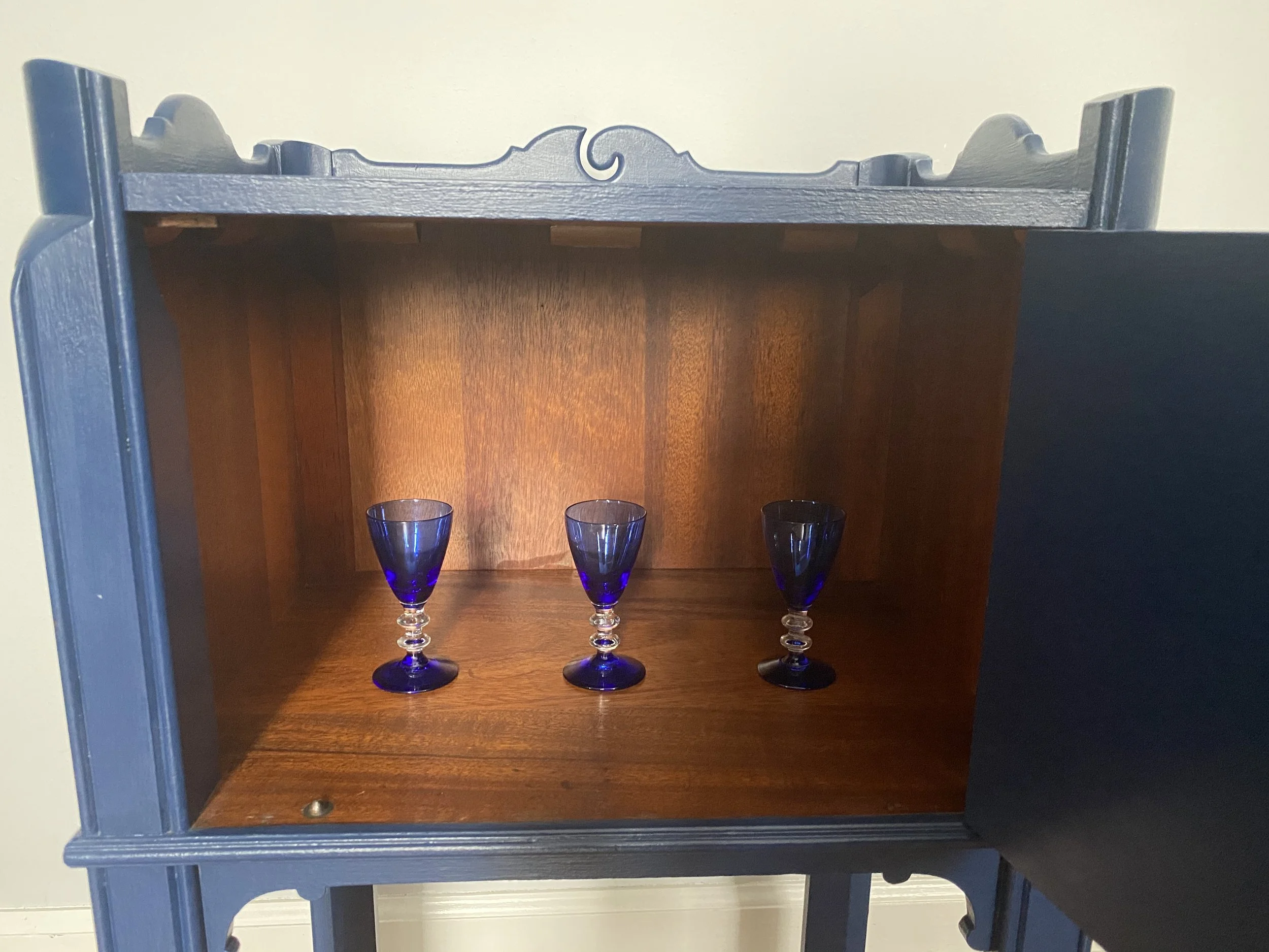 A shadow box with a dark blue frame, containing three blue wine glasses on a wooden shelf. The background inside the box is a warm wood finish.