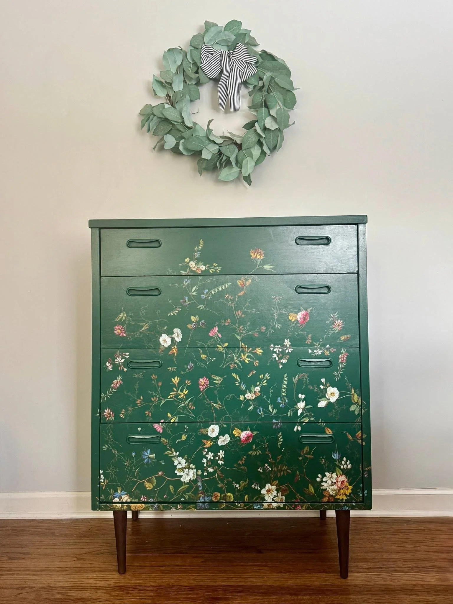 A dark green dresser with floral and leaf patterns painted on the front, standing on four wooden legs. A light-colored wall with a decorative wreath made of green leaves and a black-and-white striped bow hangs above the dresser. The floor is wooden.
