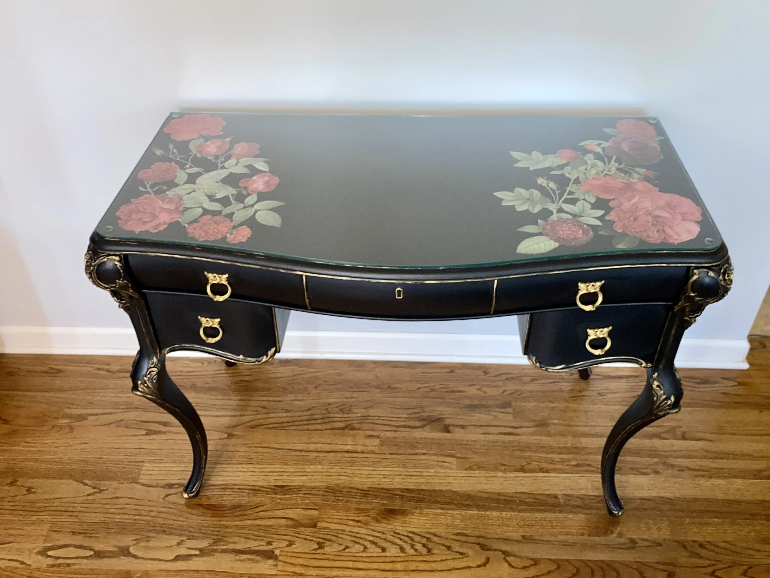 A vintage-style black table with gold embellishments and floral designs on the top surface, standing on a wooden floor against a white wall.
