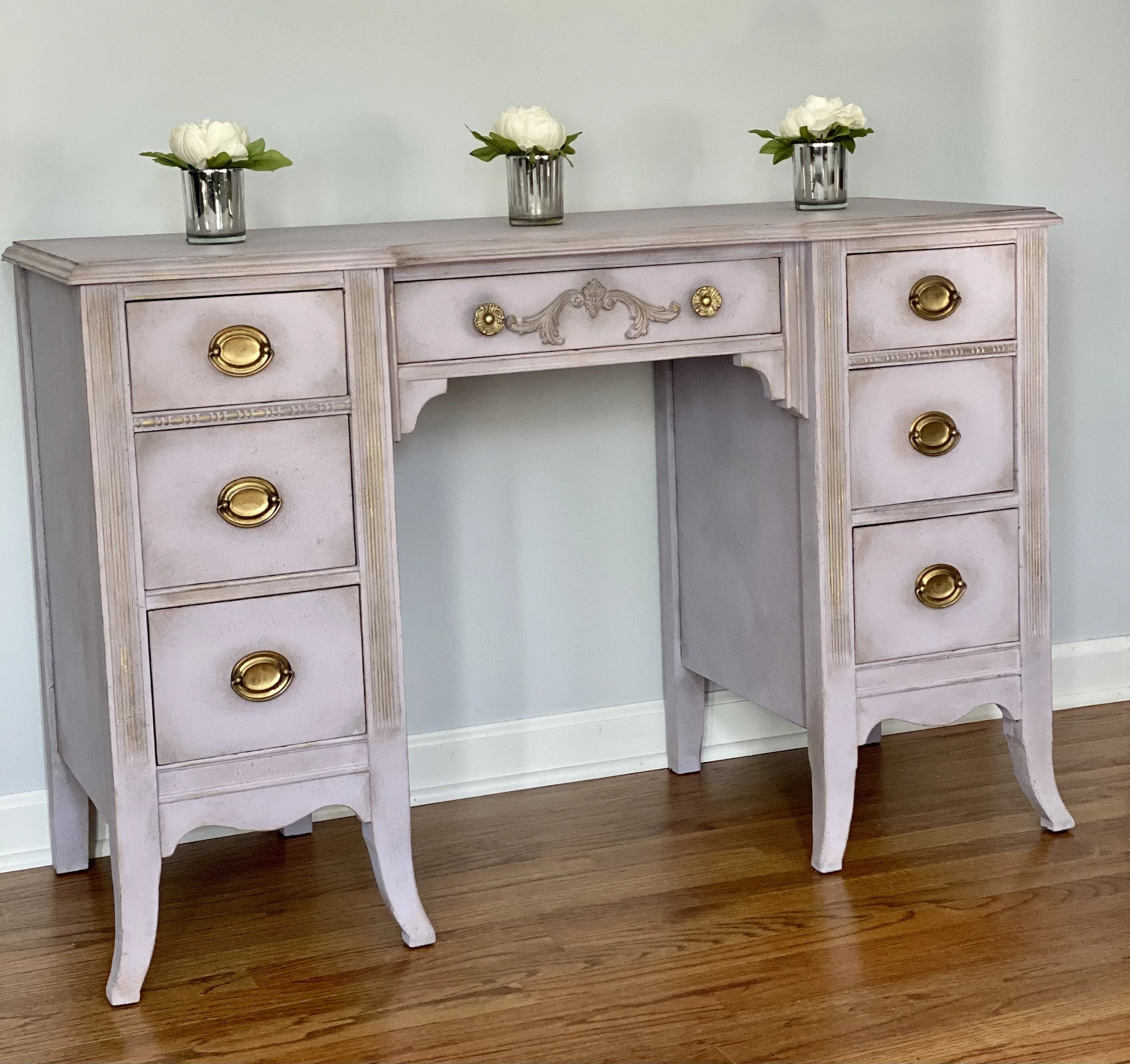 A vintage white dresser with gold knobs and carved details, placed on a wooden floor, with three white flowers in silver vases on top.