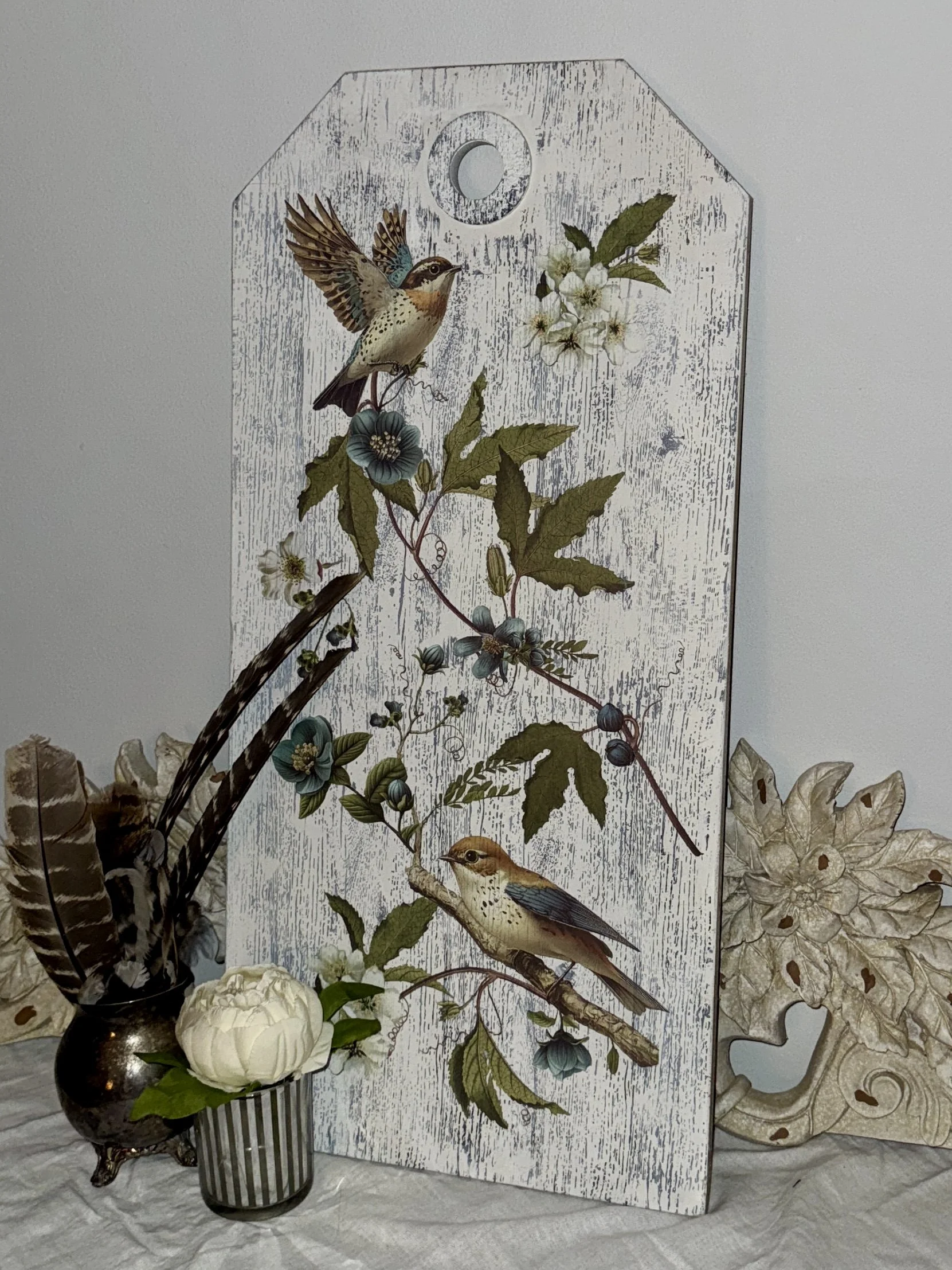 Decorative scene with a white wooden panel featuring birds and flowers, a black and white striped vase with white flowers, and a decorative stone sculpture with floral patterns on a marble surface.