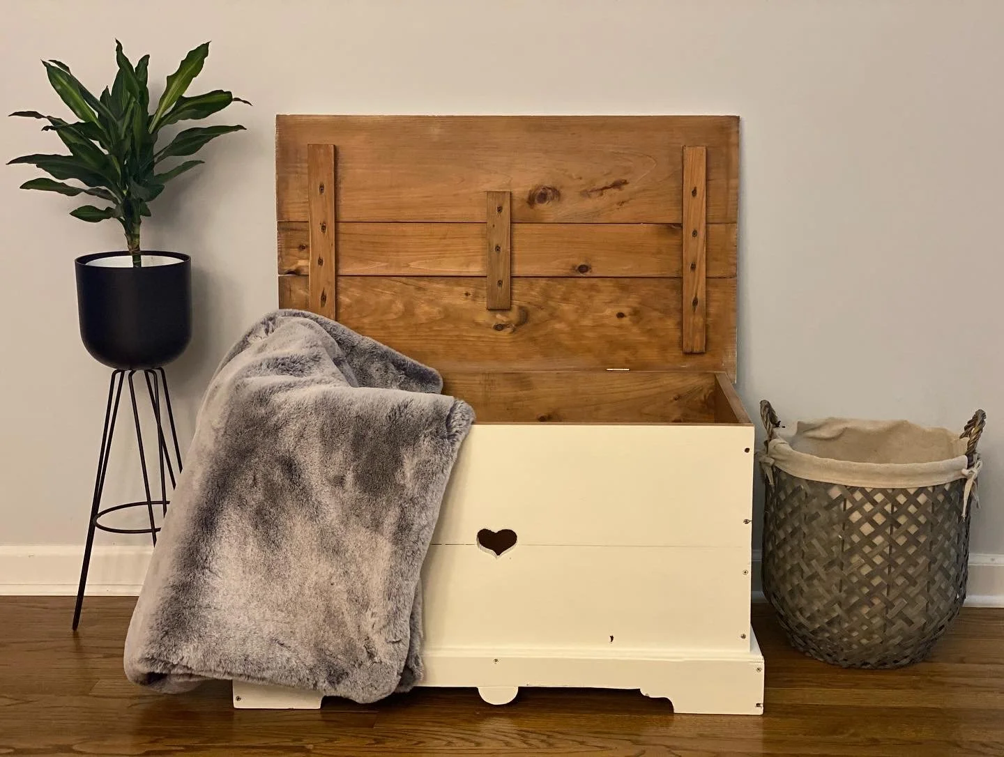 A wooden headboard with a beige storage chest in front of it, draped with a gray plush blanket. To the left, a tall black plant pot with a green leafy plant stands on a black metal stand. To the right, a woven basket with fabric lining is placed on t