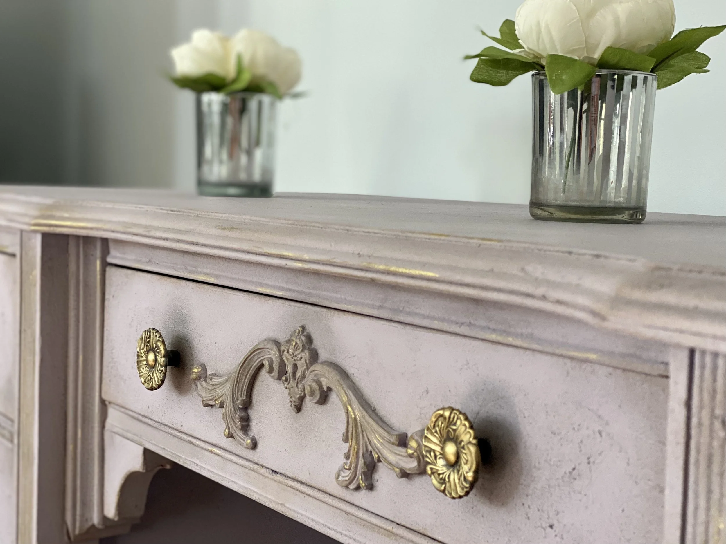 Close-up image of a decorative vintage-style dresser with ornate golden handles and a distressed white finish, topped with two glass vases containing white flowers and green leaves.