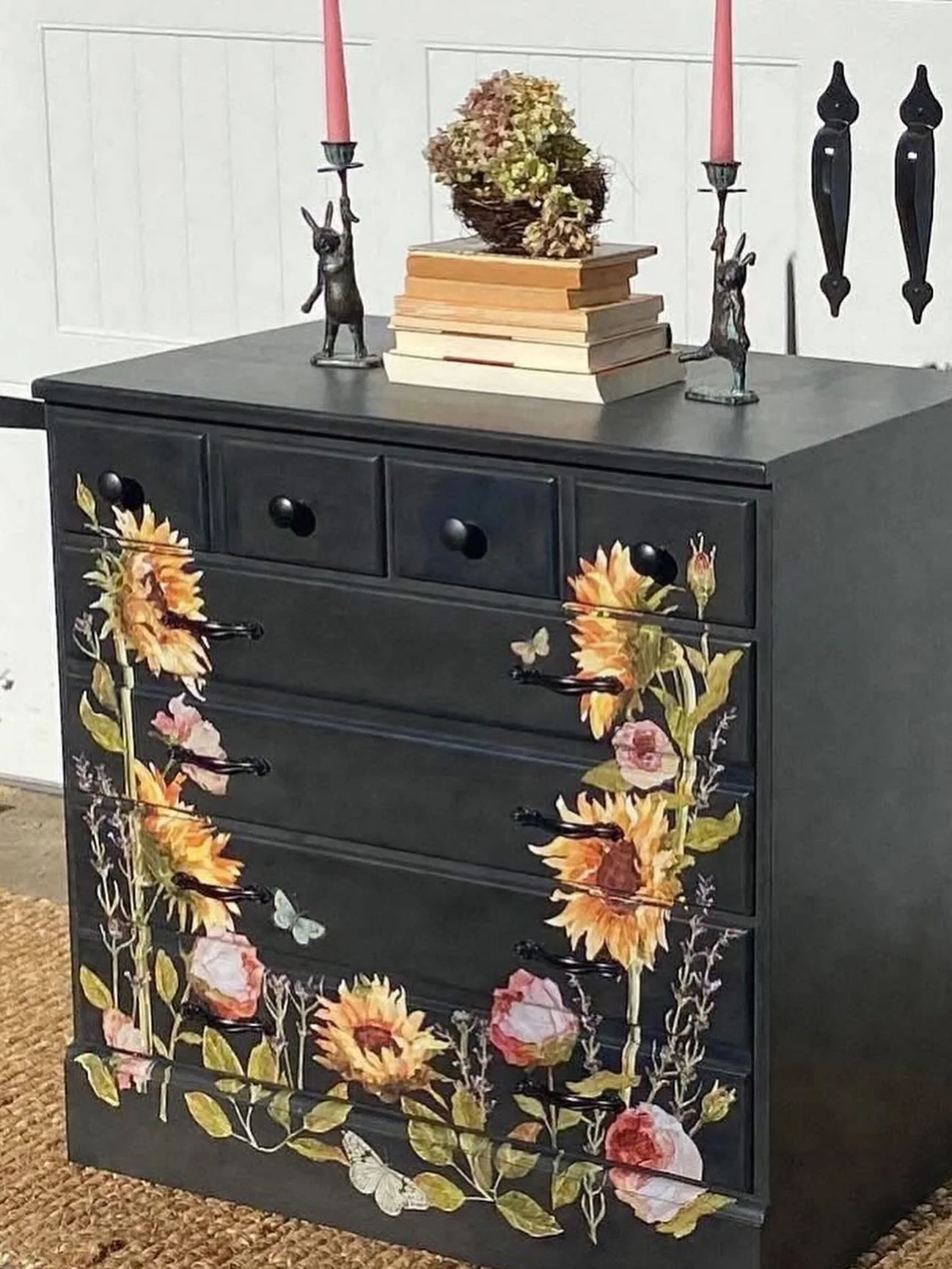 Black dresser with sunflower and pink flower floral design on the front, topped with a stack of books, a dried floral arrangement, and two candle holders with pink candles.