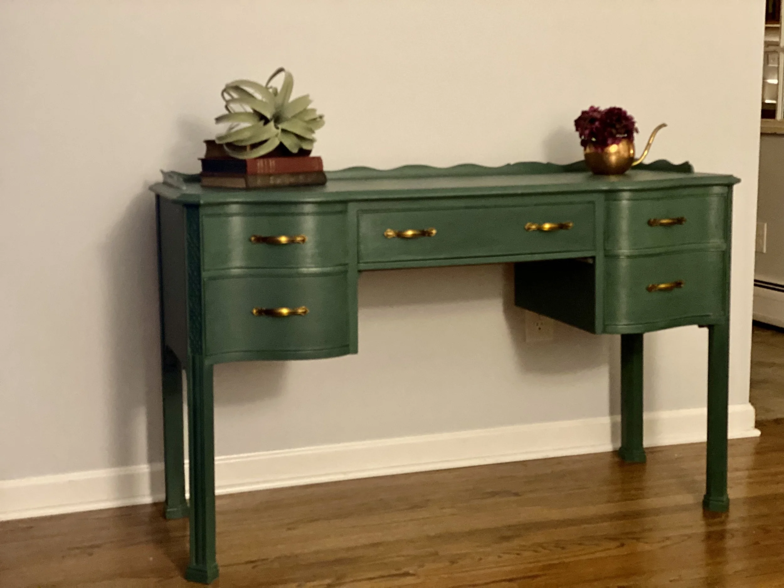 A green vintage-style sideboard with golden handles, decorated with a potted plant and a small gold watering can filled with red flowers, against a plain white wall with a hardwood floor.