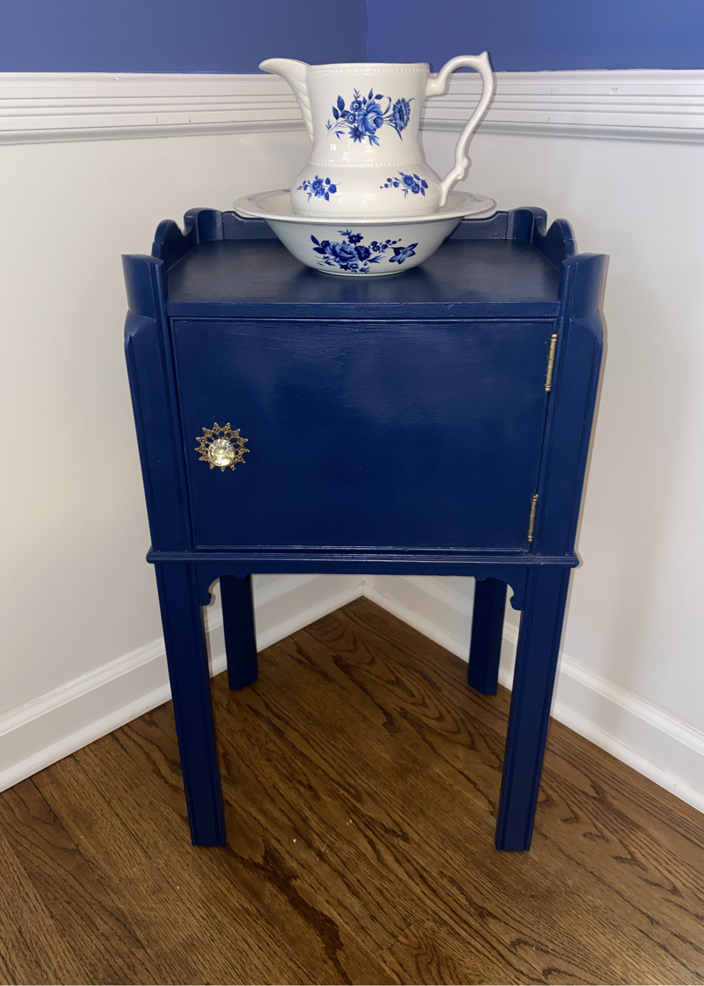 A small, blue-painted wooden table with a decorative gold and pearl knob on the door, holding a white porcelain pitcher and bowl with blue floral patterns, against a wall with white trim and a blue wall above.