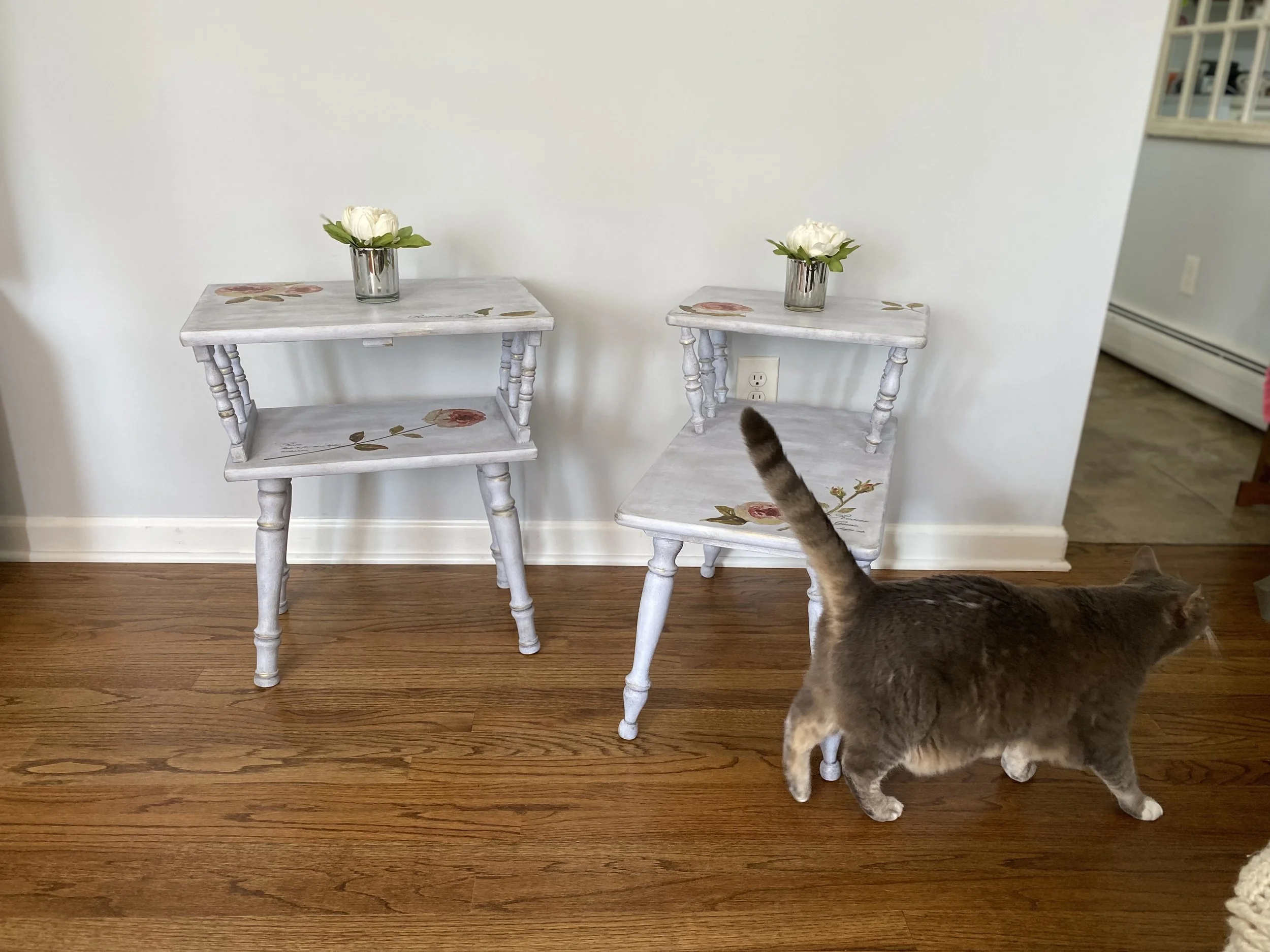 Two small white tables with floral designs leaning against a plain white wall. Each table has a small glass vase with a white flower and green leaves. A gray and brown cat is partially visible in the bottom right corner, walking past the tables. The 