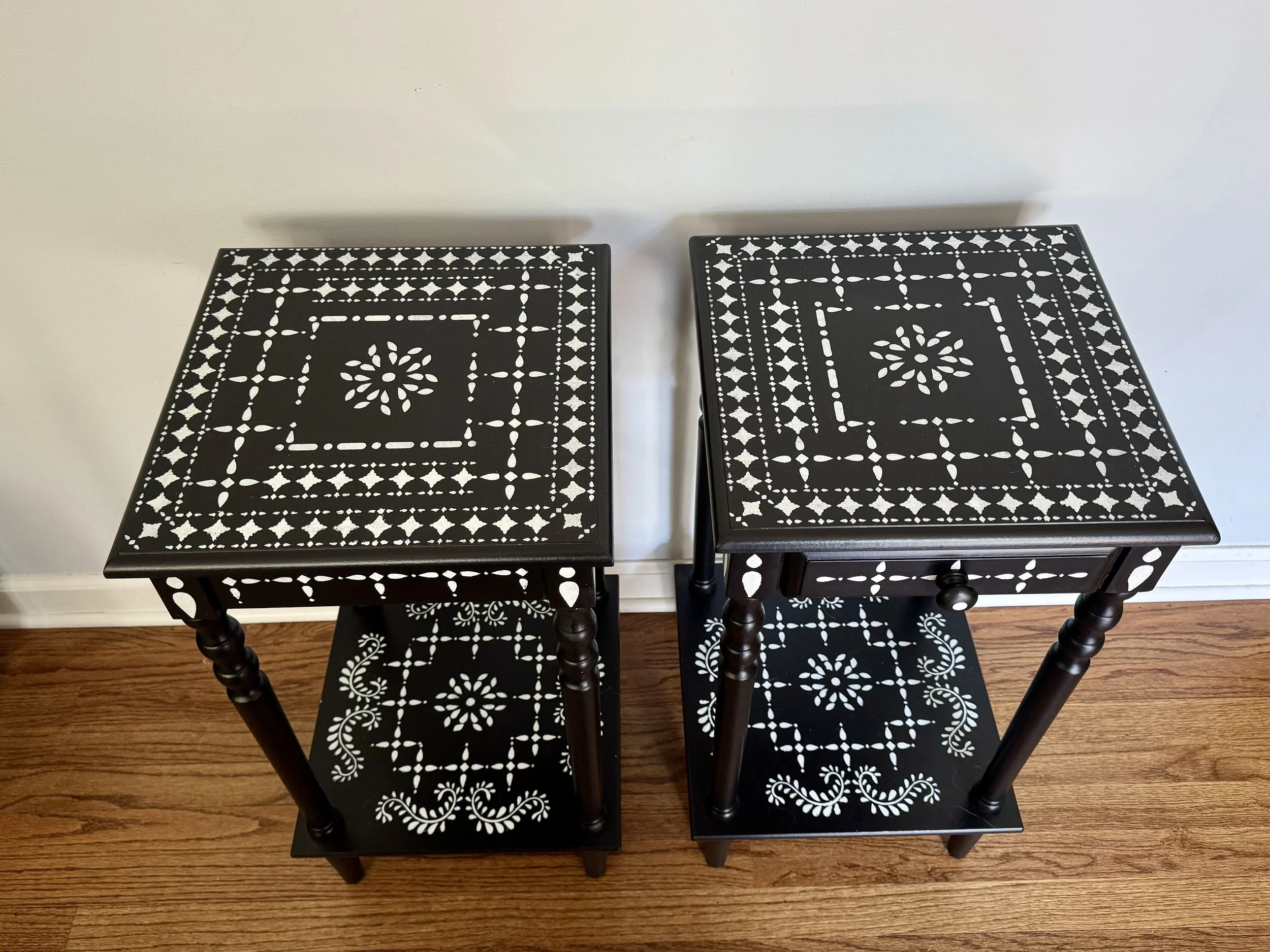Two black wooden chairs with white floral and geometric patterns on the seats and backs, positioned on a wooden floor against a plain light-colored wall.