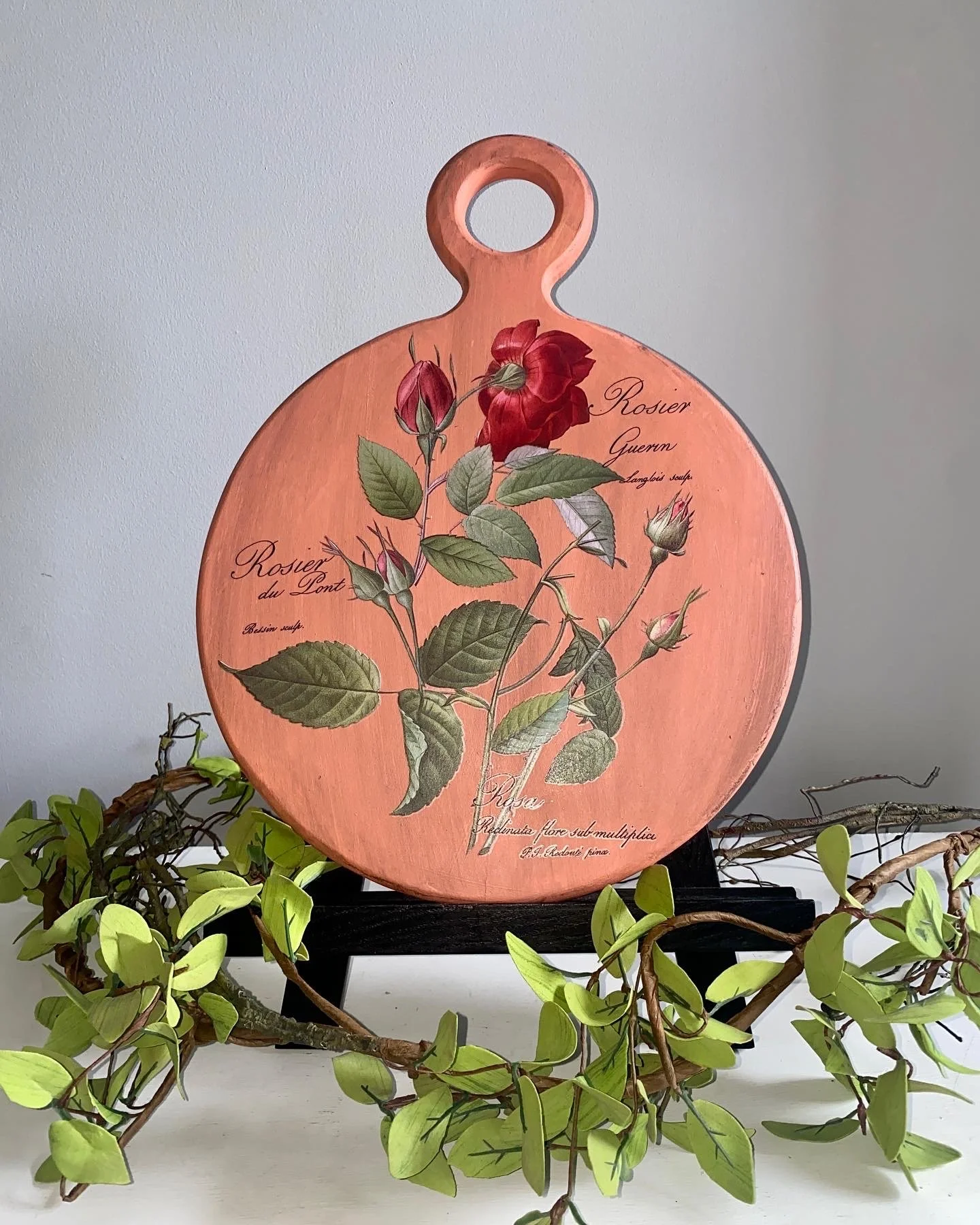 A decorative wooden cutting board with botanical illustrations of roses, including buds and leaves, labeled in Latin and French, resting on a black stand. Green vine-like plants surround the base.