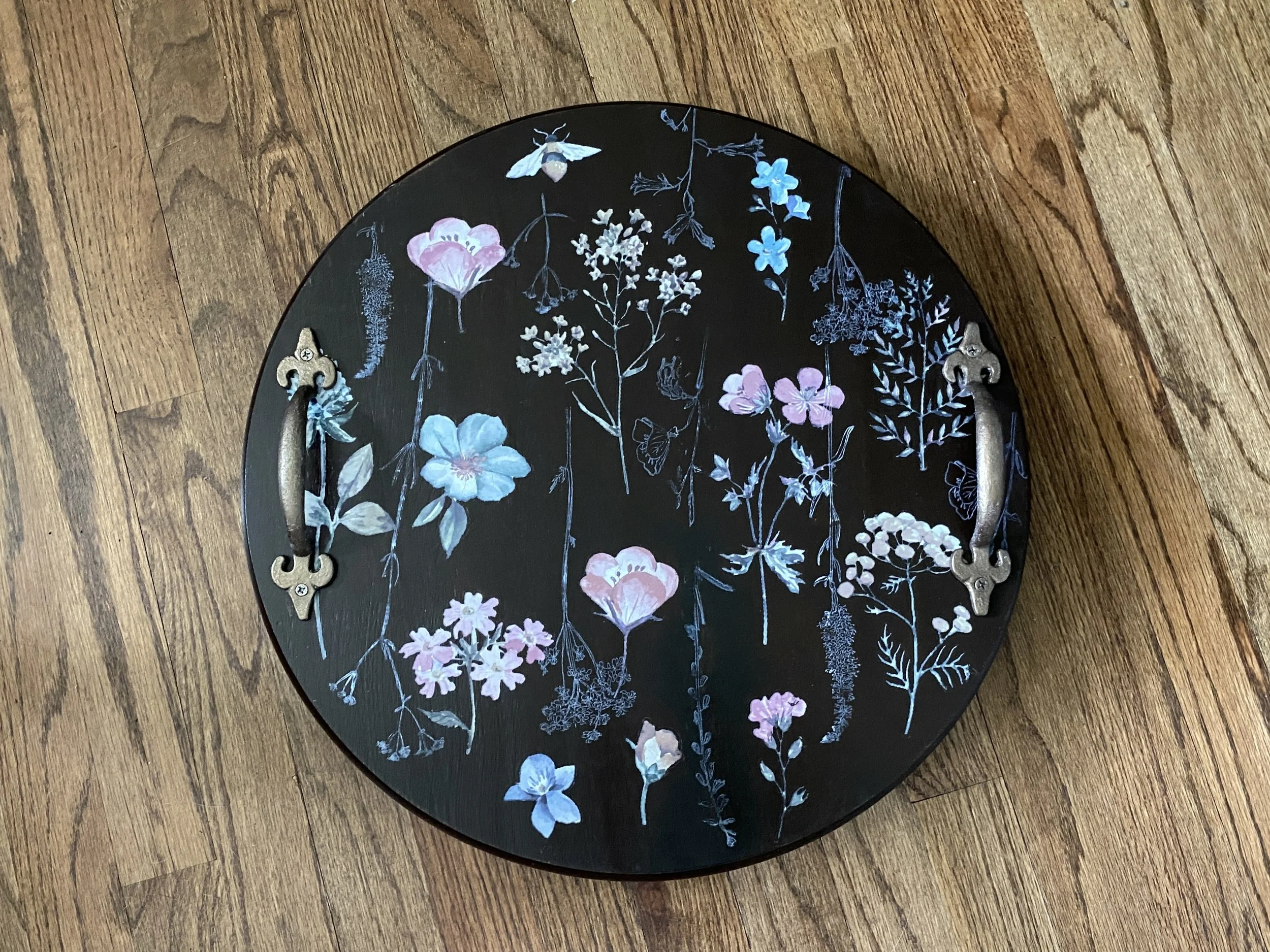 A black round tray with a floral pattern of pink, blue, and white flowers, and a white bee, placed on a wooden floor.