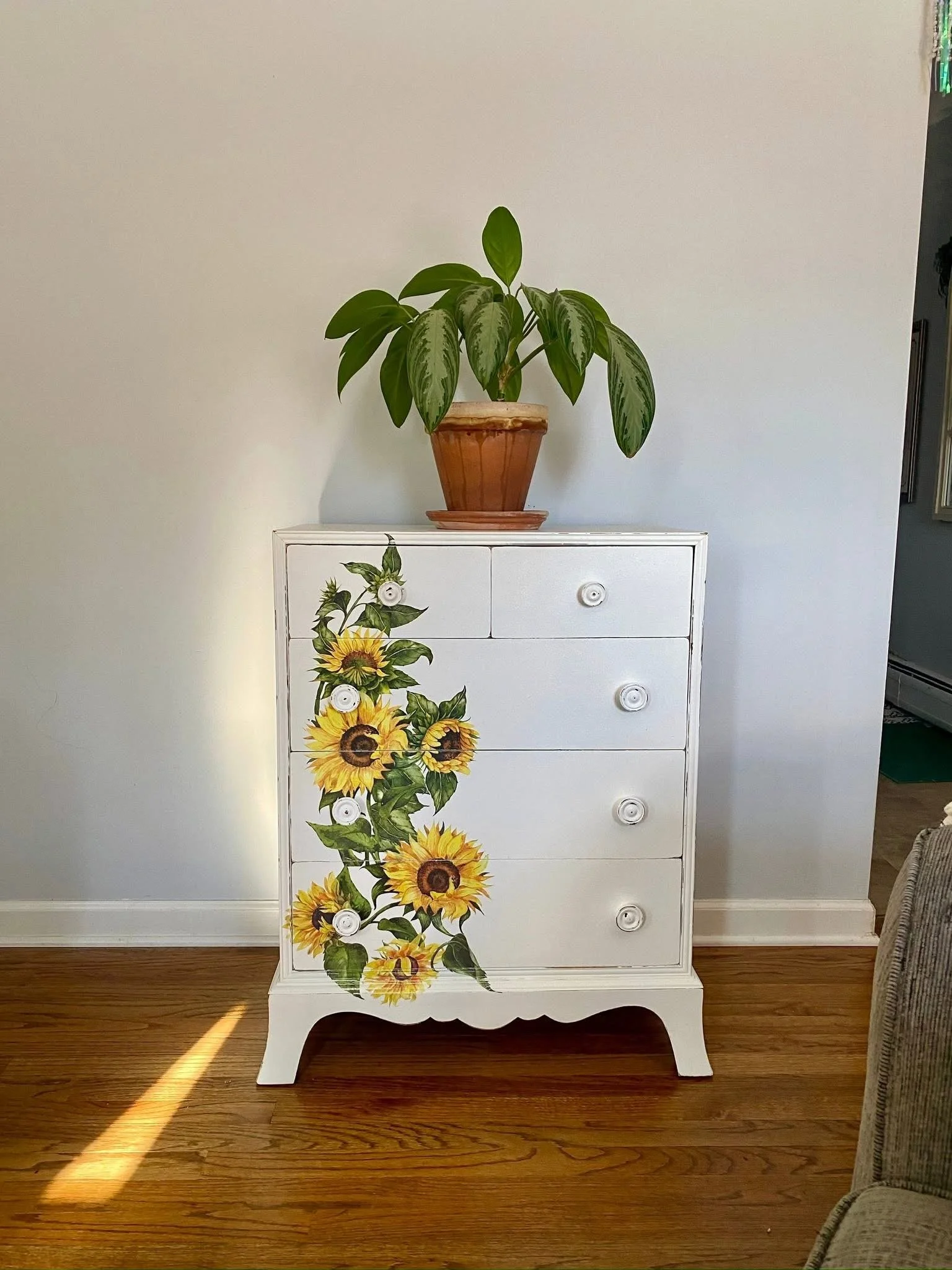A white dresser with a sunflower and vine painted on its front, featuring six drawers with white knobs. A potted plant with green leaves is placed on top of the dresser against a plain wall.