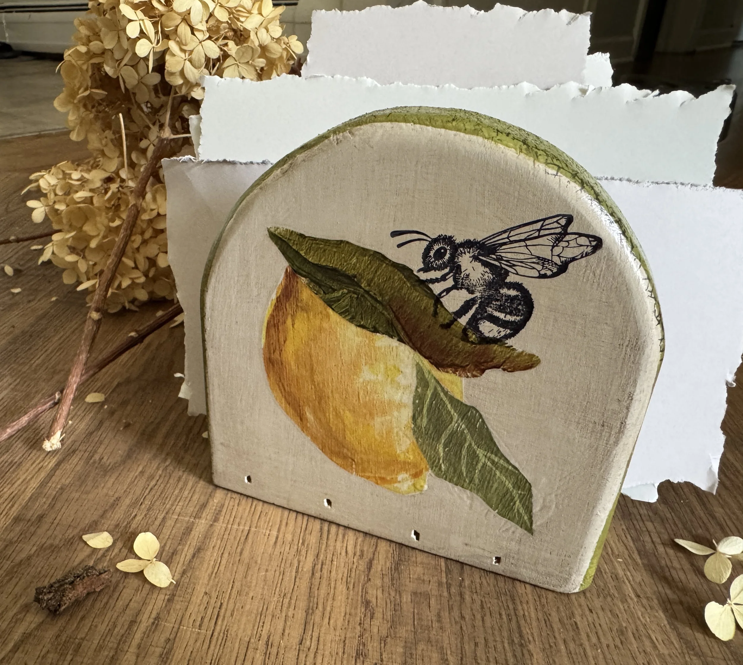 A wooden surface with dried hydrangea flowers and a piece of torn white paper or cardboard with a bee illustration behind a carving of a leaf.