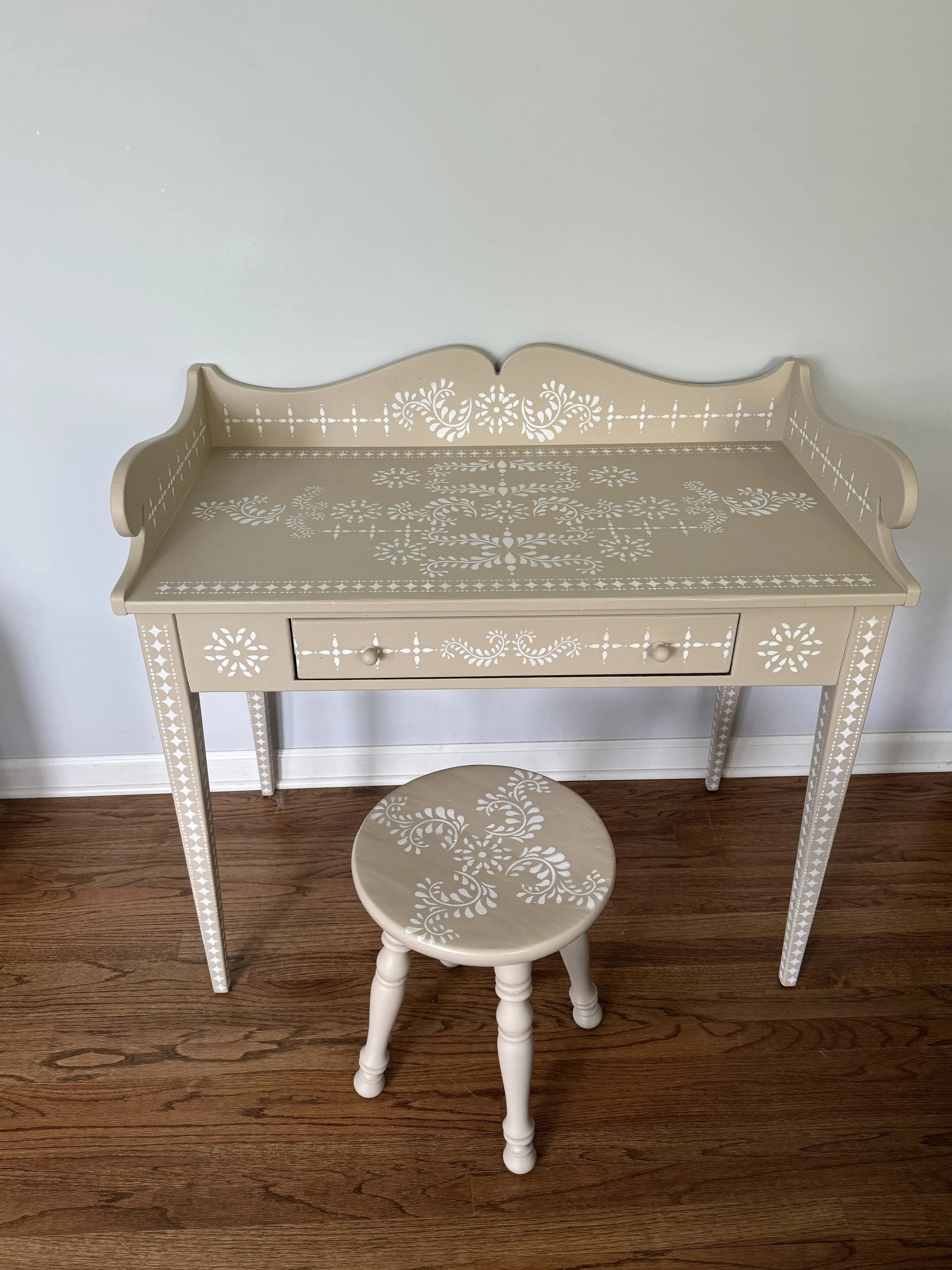 A white wooden vanity table with a matching stool, both decorated with intricate star and floral patterns, placed on a hardwood floor against a plain wall.