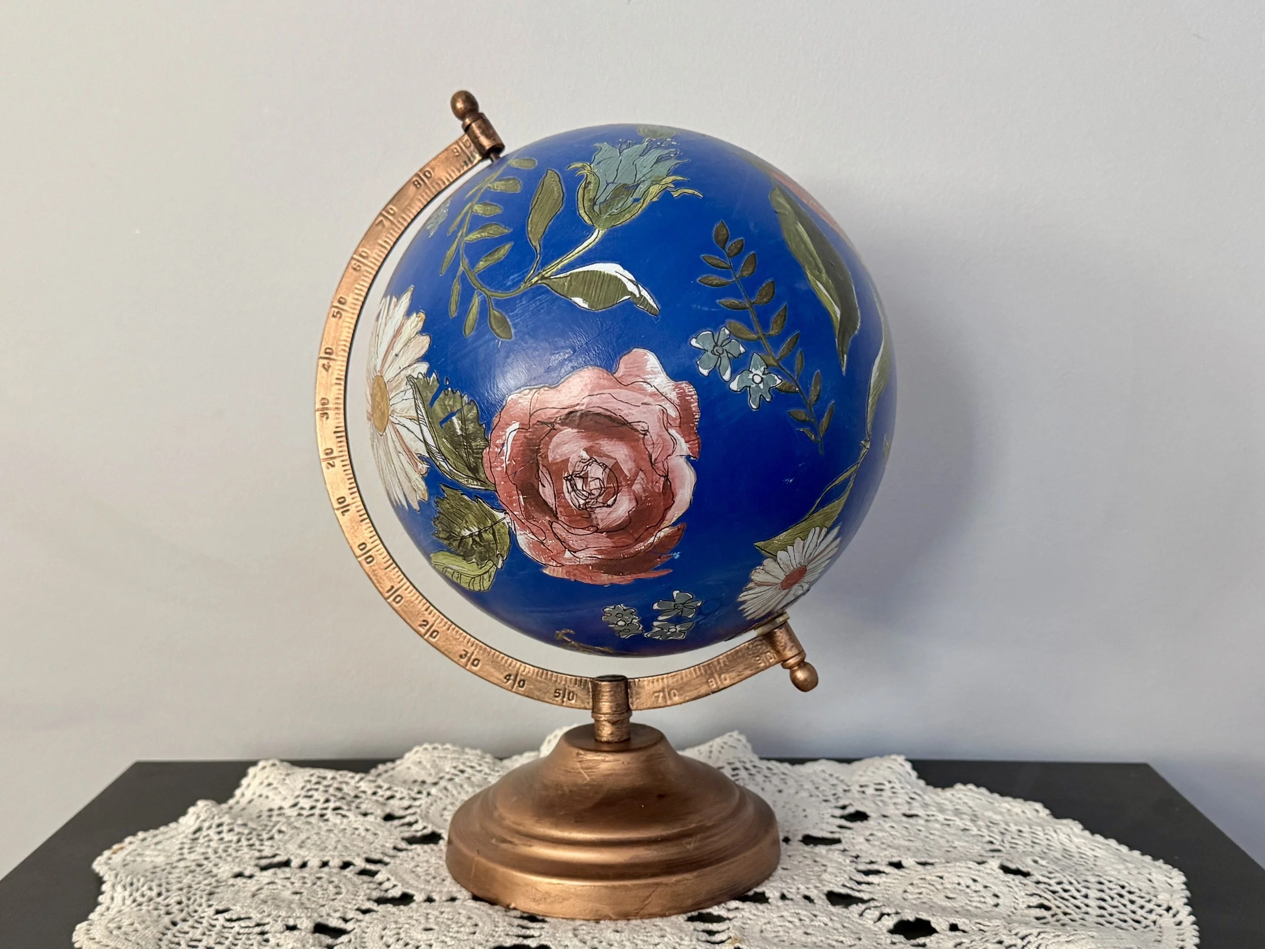A decorative globe with a painted floral design, featuring pink, white, and blue flowers with green leaves, mounted on a copper-colored stand, placed on a white lace doily against a plain background.