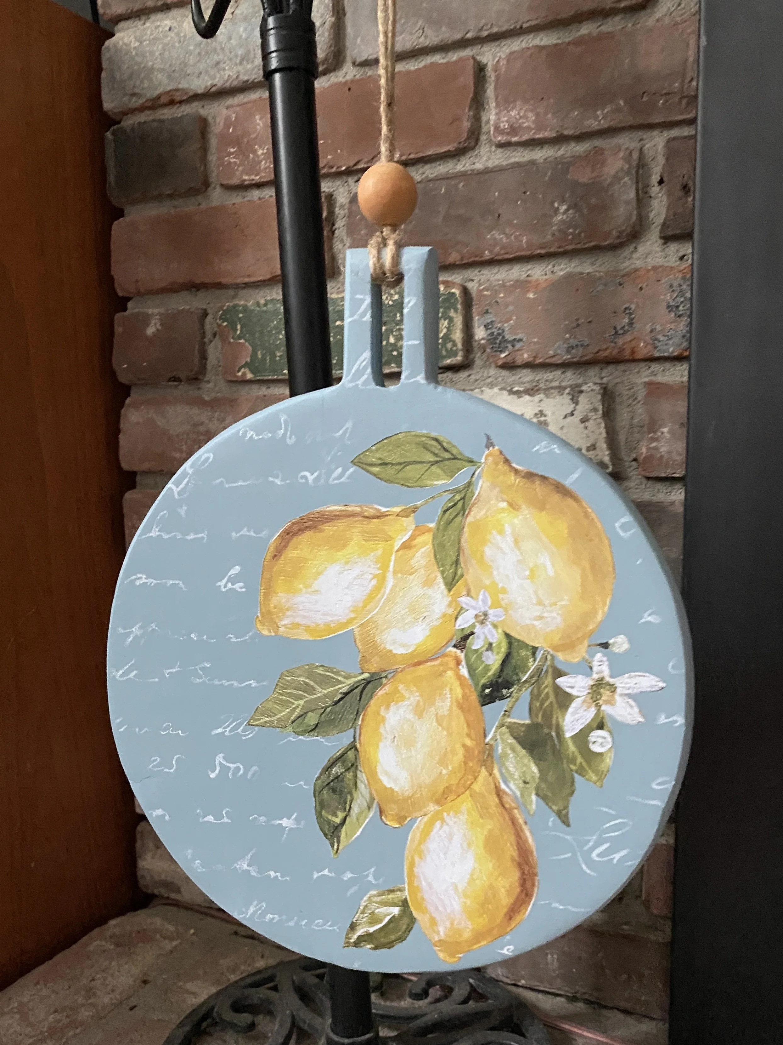 A decorative round serving tray with a painted lemon design hanging from a hook on a brick wall, with a black vertical pole to the left and a wooden surface to the left side of the image.