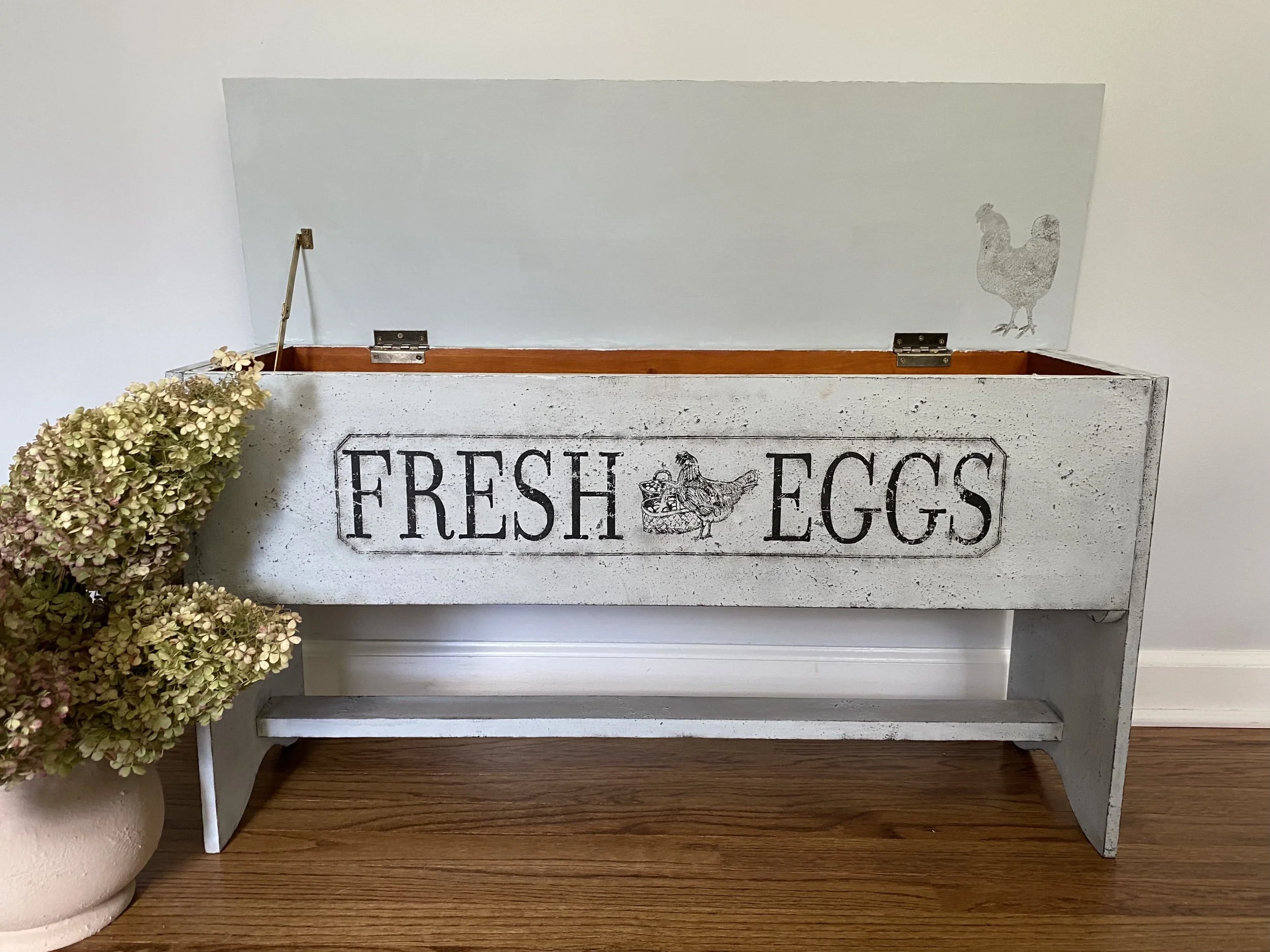 White wooden hatch with black printed sign that says 'Fresh Eggs', a black and white hen illustration, and a gray chicken silhouette. A beige vase with dried hydrangea flowers is placed beside the hatch on a wooden surface.