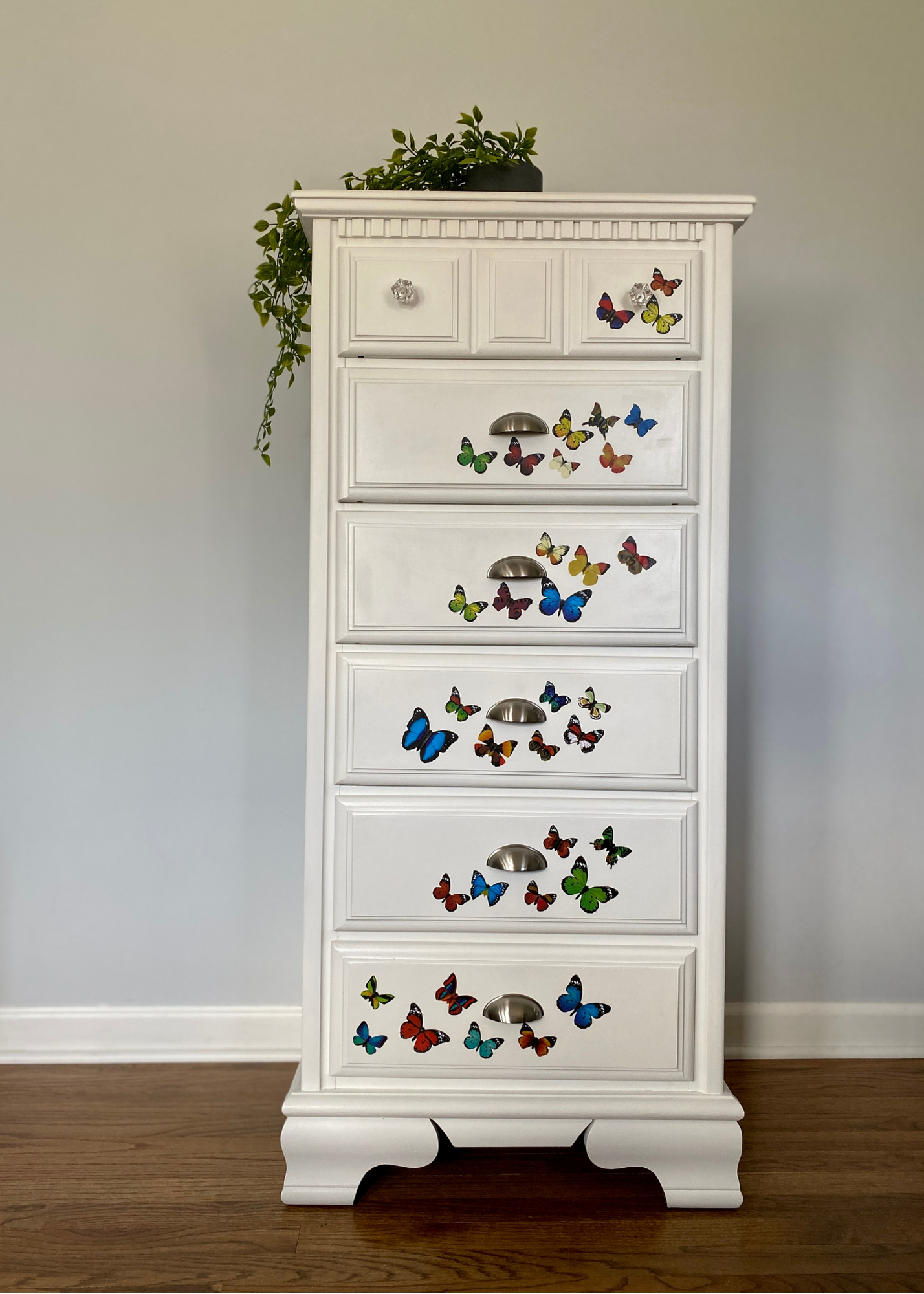 A white wooden dresser decorated with colorful butterfly stickers in various sizes and colors. There is a leafy green plant on top of the dresser, hanging slightly over the edge. The dresser has four drawers with metal handles and is situated against