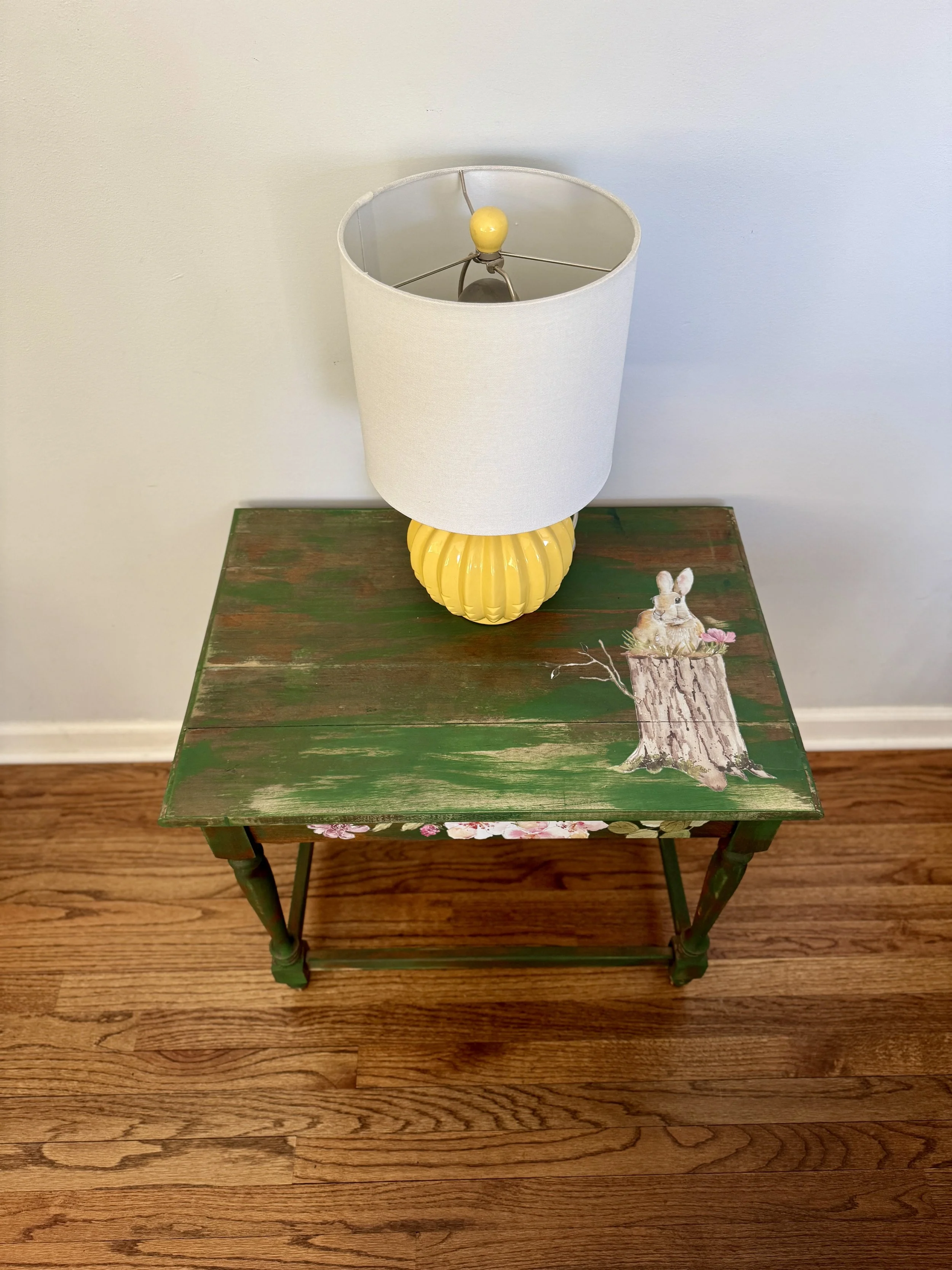 A wooden table painted green with pink flowers and a bunny illustration, against hardwood flooring and a white wall, with a lamp on top.