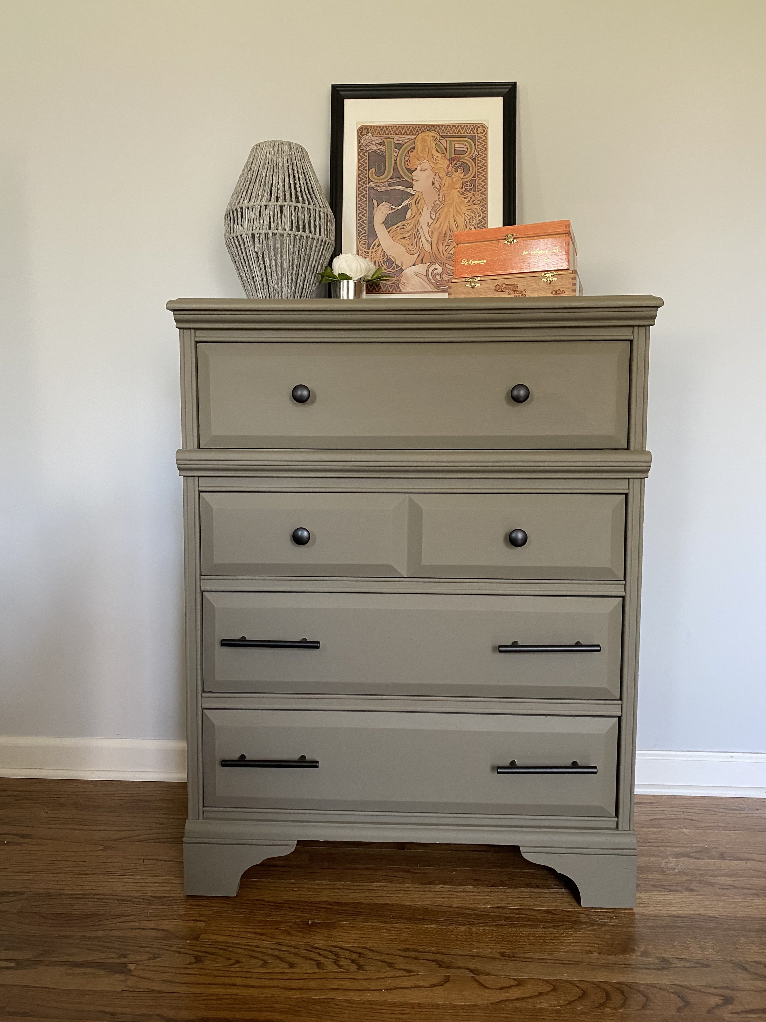 A beige wooden dresser with six drawers, two with black handles, positioned on a hardwood floor against a plain white wall. On top are decorative items including a large gray woven vase, a framed artwork of a woman with long flowing hair, a small whi
