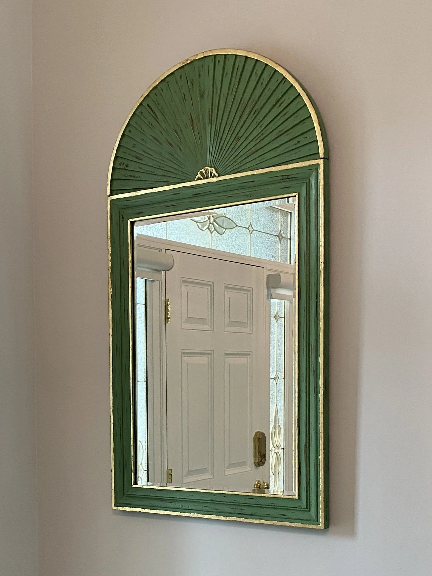 A rectangular mirror with a green and gold frame hanging on a beige wall, reflecting a white door with sidelights and stained glass panels.
