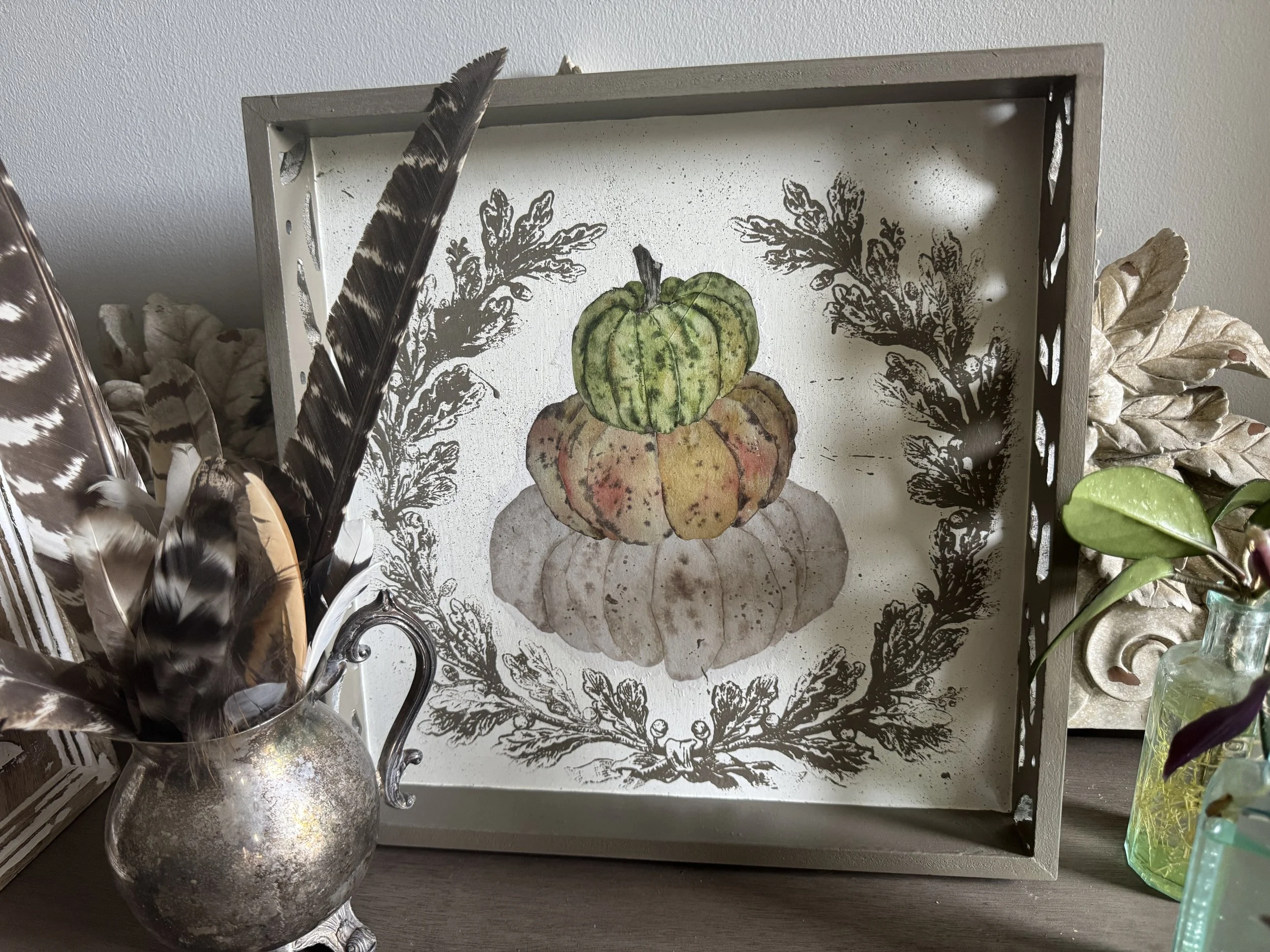 Decorative tray with a pumpkin illustration, surrounded by feathers, dried leaves, and a glass container.