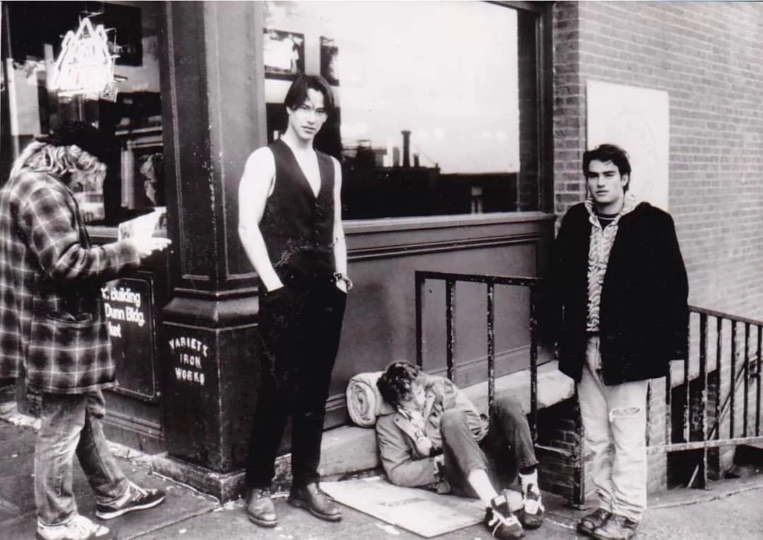 Three young people outside a building, one sitting on the ground leaning against the wall, two standing nearby, one looking at their phone, and another looking at the camera, with a window and brick wall in the background.
