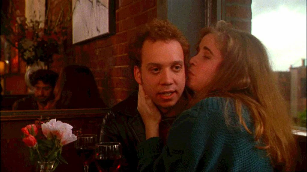 A man with curly hair and a woman with long hair are in a romantic pose in a restaurant. The woman is leaning in to kiss the man's cheek, and he has a surprised or intense expression. There are two glasses of red wine and a small vase with pink and white flowers on the table. In the background, a couple is sitting at another table near a brick wall.