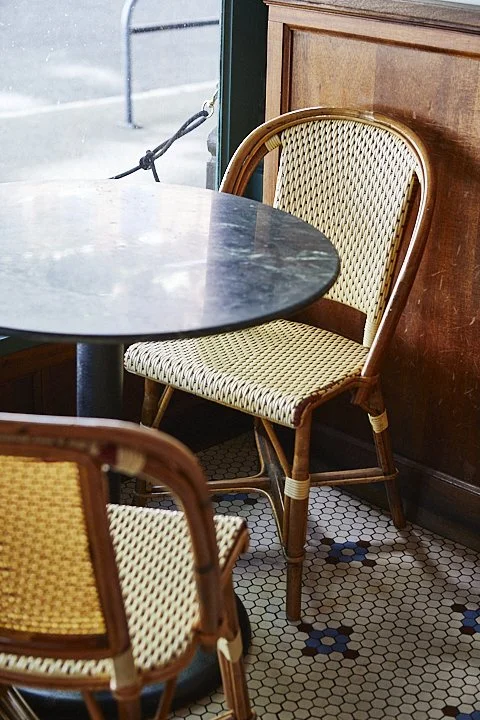 Inside a cafe, a round marble table with wicker chairs, one with a beige and brown woven pattern, near a wooden wall and large window.