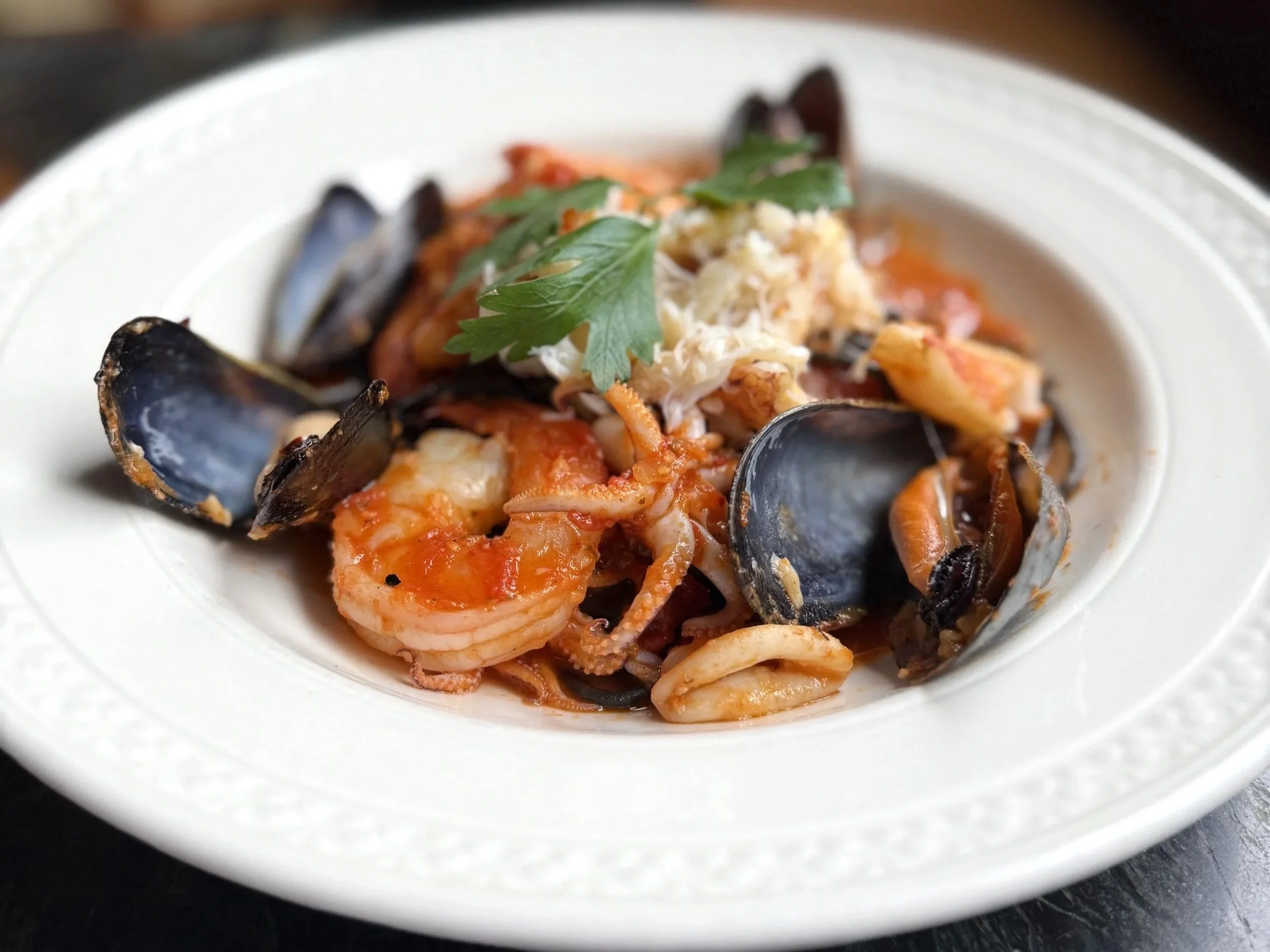 Seafood dish with mussels, shrimp, and chunks of fish garnished with a sprig of parsley on a white plate.