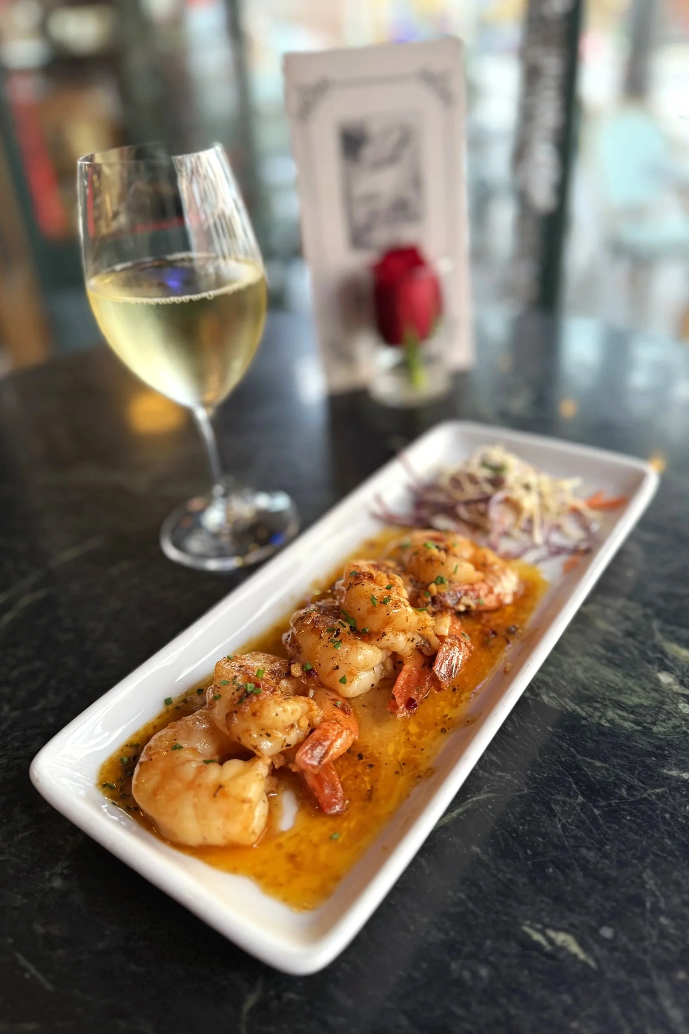 A white rectangular plate of cooked shrimp in a sauce with herbs, served with a side of coleslaw, a glass of white wine, and a blurred background of a restaurant setting with a rose and a menu on the table.