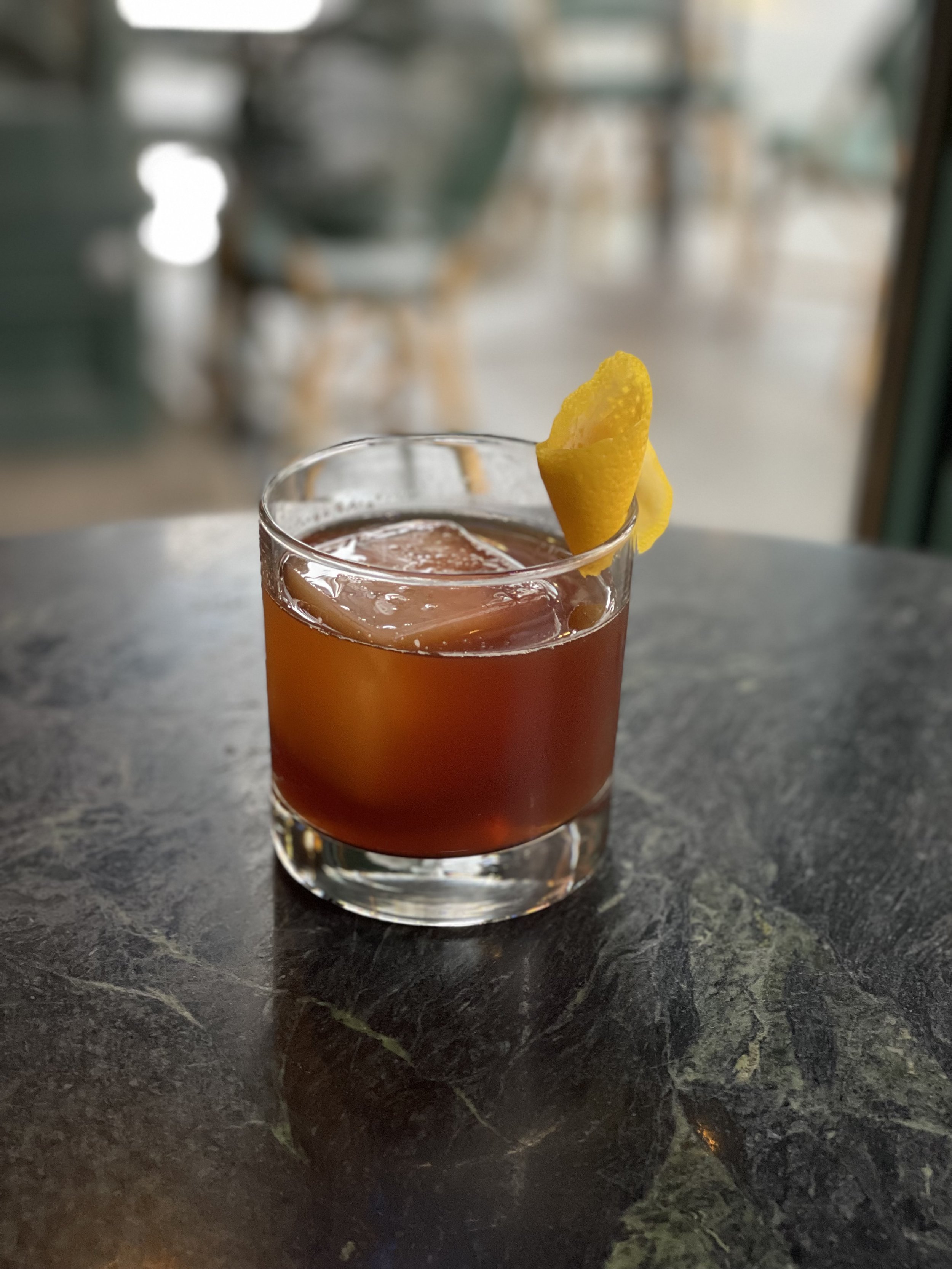 A cocktail glass with dark reddish-brown liquid and a lemon peel garnish, sitting on a dark marble table.