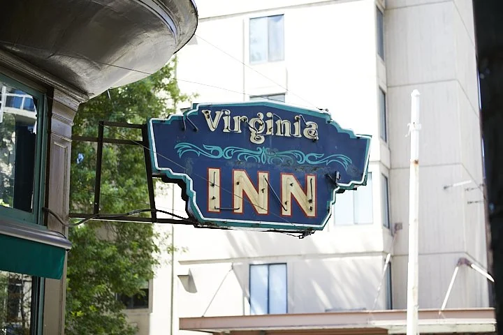 Neon sign for the Virginia Inn hotel in an urban area with tall buildings and trees.