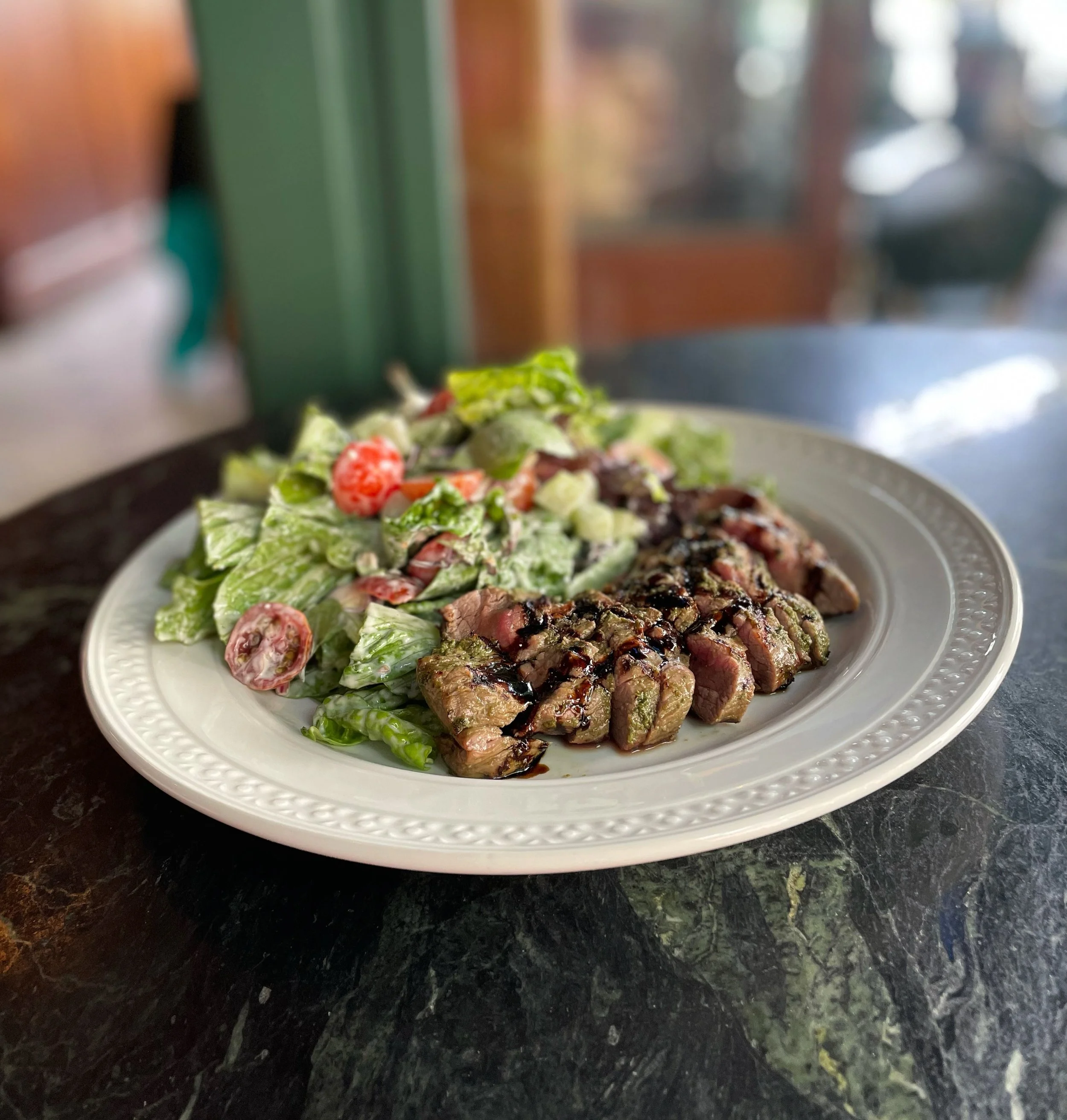 A white plate with a side salad and sliced cooked steak drizzled with balsamic glaze on a dark tabletop, with a blurred background.