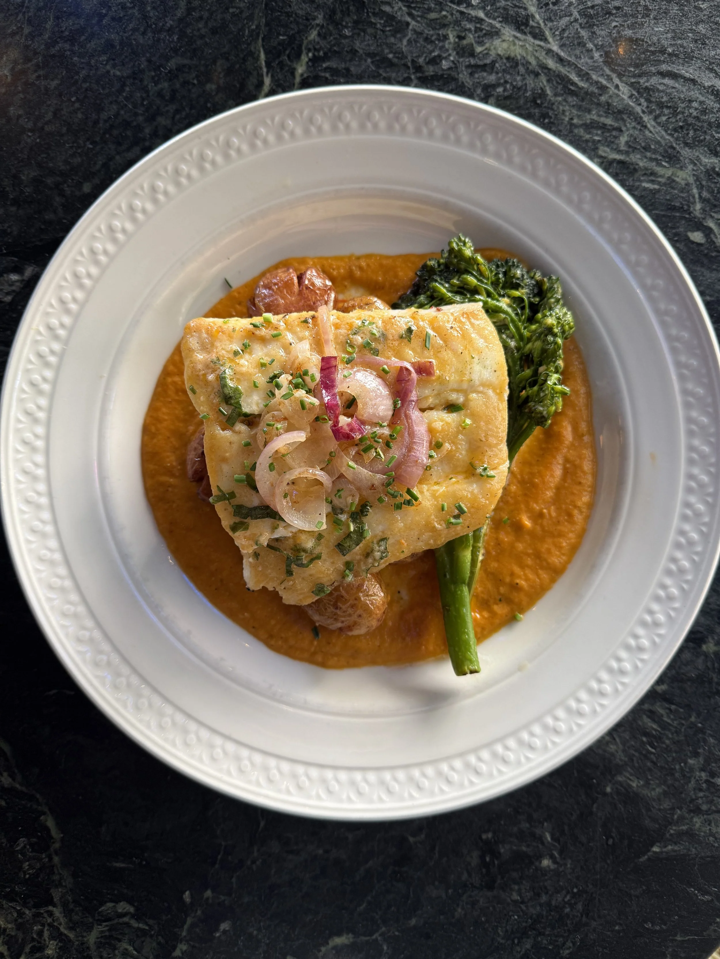 A plate of fish topped with chopped onions and chives, served on a bed of orange sauce with broccoli on the side.