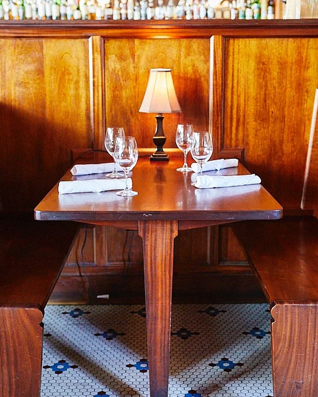 A wooden restaurant booth with a set dining table, four wine glasses, four white napkins, and a small table lamp on a wooden wall.