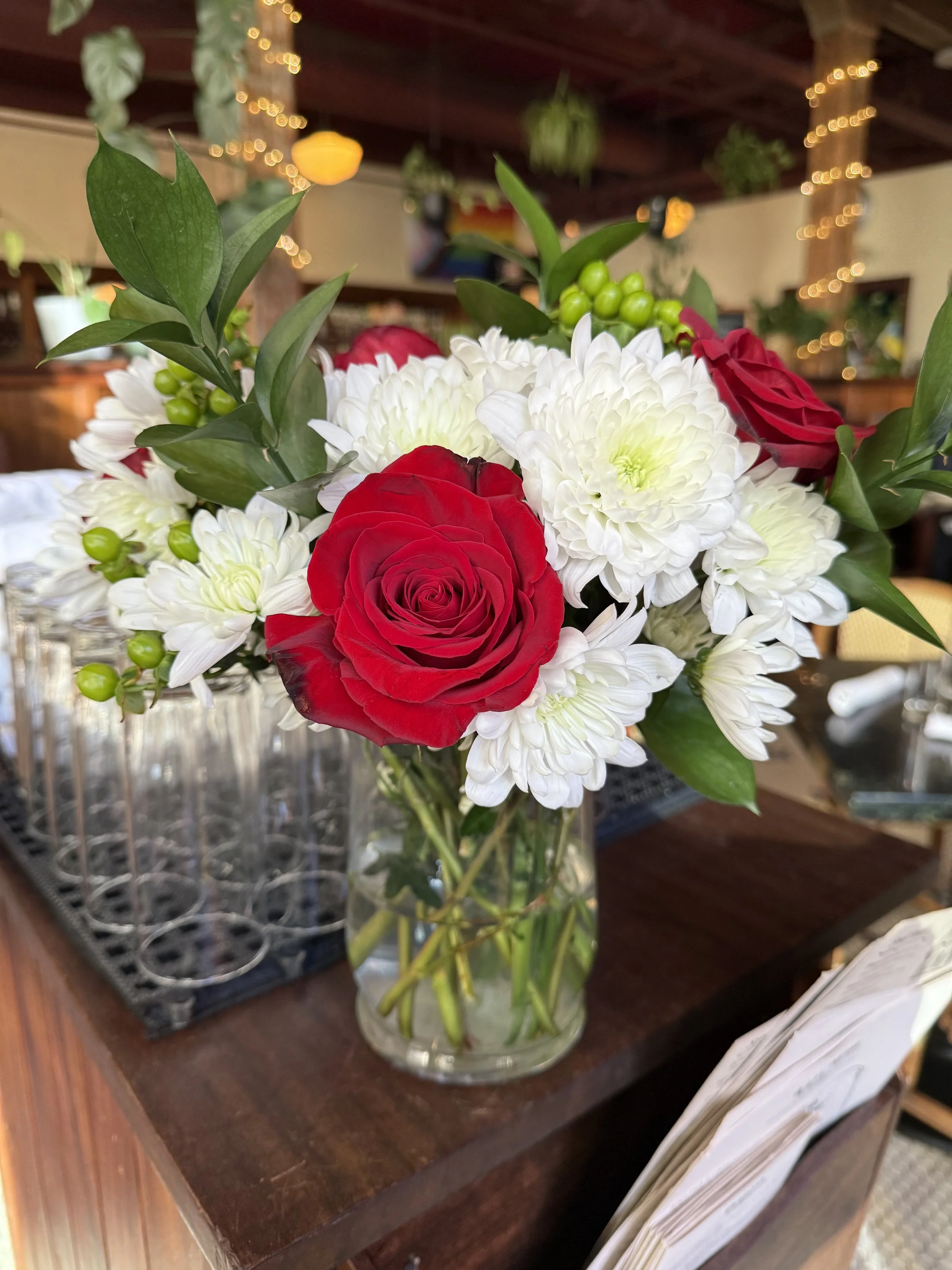 A bouquet of red roses white chrysanthemums and green berries in a clear glass vase on a wooden surface, with a background of warm lighting and hanging plants.