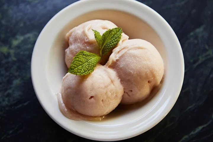 Three scoops of pink ice cream garnished with fresh mint leaves in a white bowl on a dark surface.