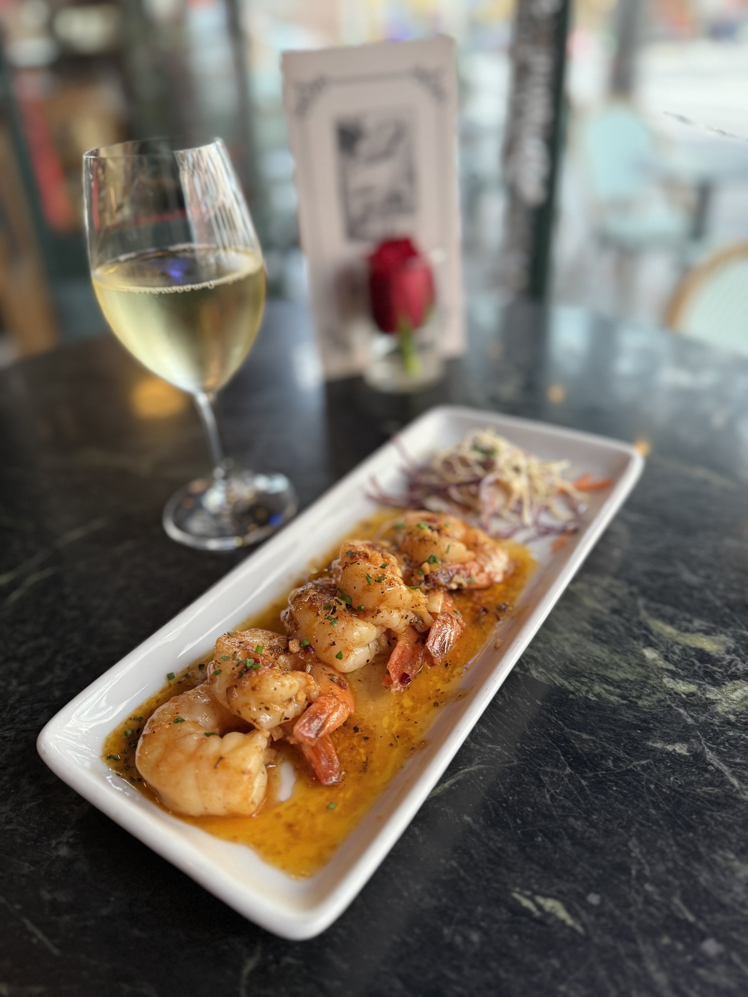 A wine glass filled with white wine, a rectangular dish with cooked shrimp in orange sauce, and a small serving of coleslaw on a dark table, with a blurred background including a menu and a rose in a glass.