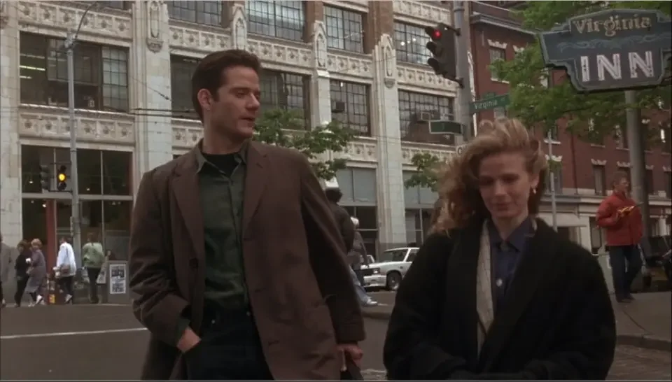 A man and woman walking on a city street with buildings, cars, and pedestrians in the background. The man has dark hair and is wearing a brown jacket, while the woman has blonde hair and is wearing a dark coat.