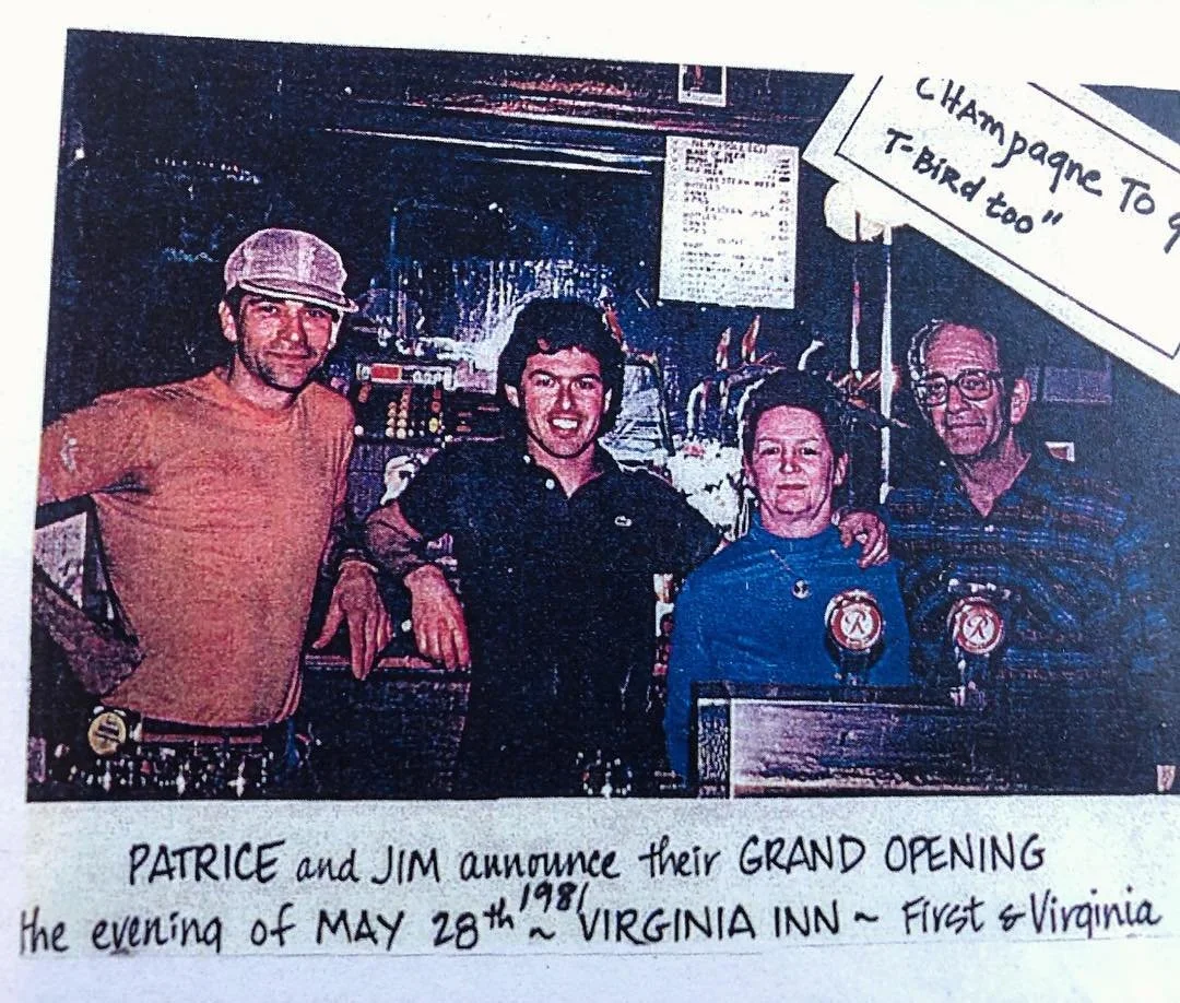 Four people standing behind a bar, celebrating a grand opening. The person on the far left is wearing a beige cap and orange shirt, the second person is in a dark shirt, the third person is in a blue shirt, and the person on the far right is wearing glasses and a checkered shirt. There is a sign in the corner that reads 'Champagne To q T-Bird too'. Handwritten text at the bottom says 'Patrice and Jim announce their grand opening the evening of May 28th, 1981 Virginia Inn - First & Virginia'.