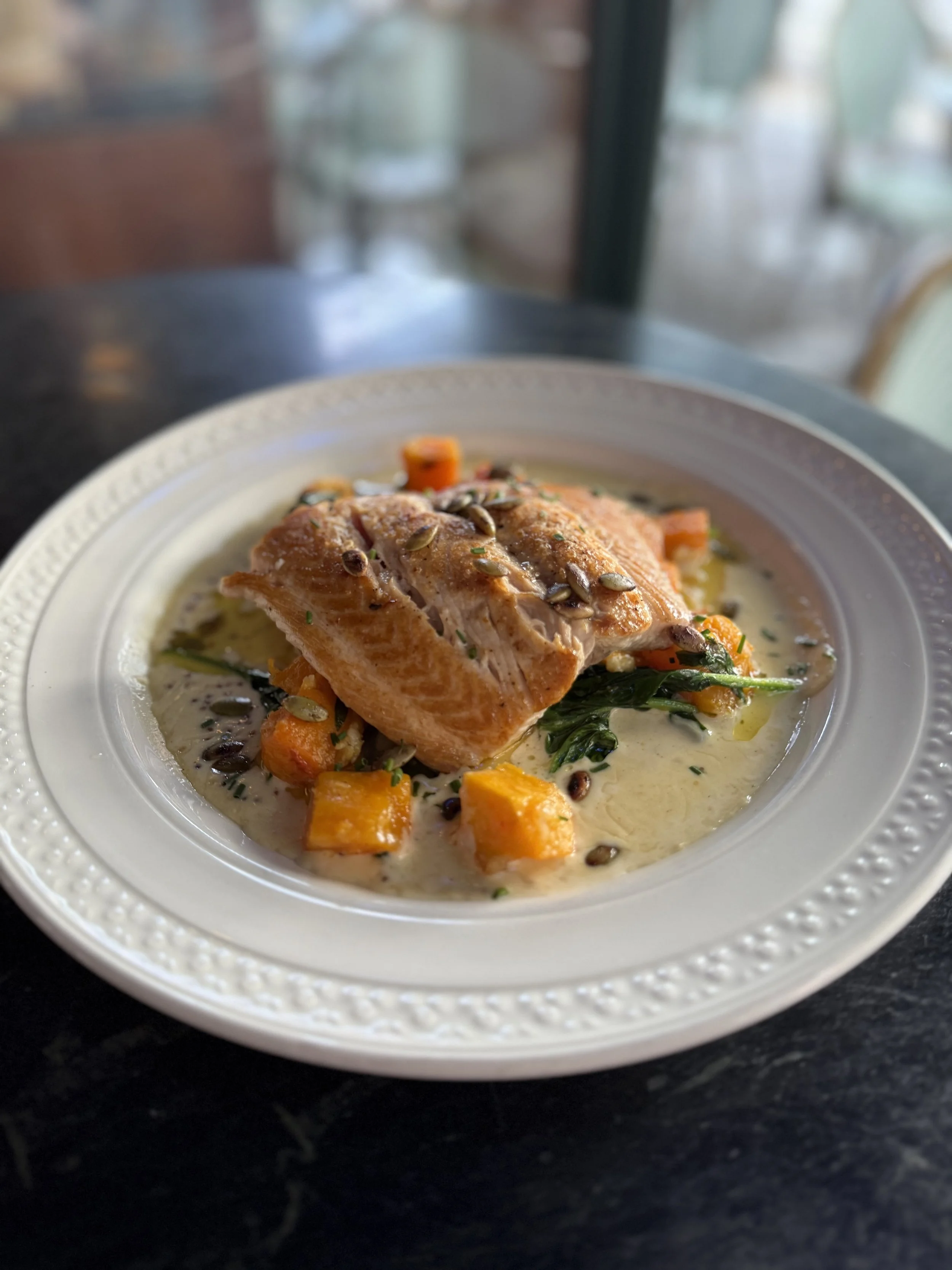 Plate of cooked salmon garnished with sunflower seeds, served with roasted sweet potatoes, spinach, and a creamy sauce.