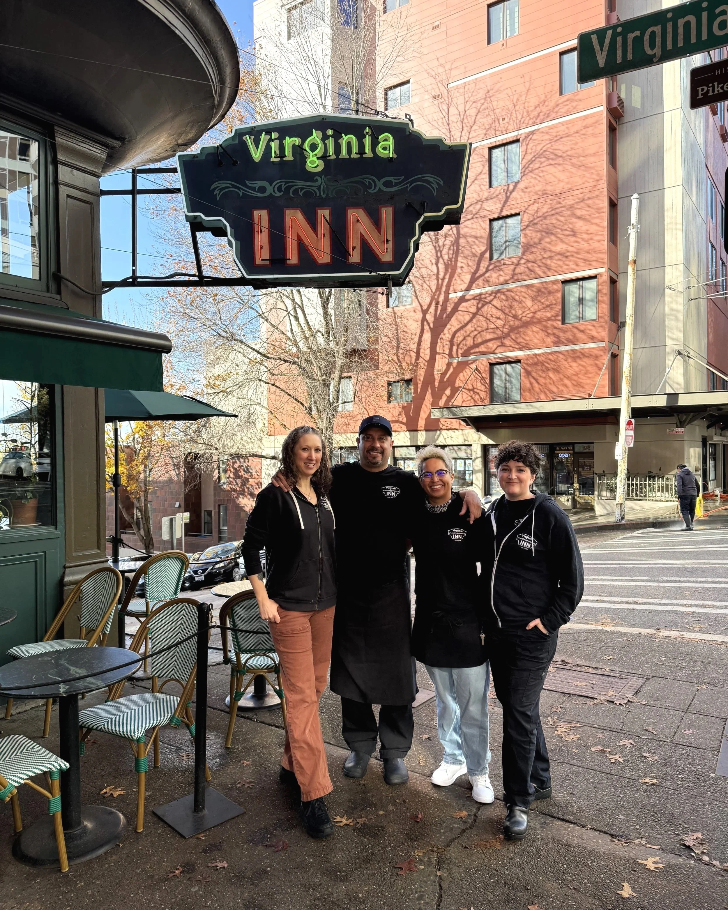 Four people standing outside the Virginia Inn, a neon-lit sign above them displaying "Virginia INN." The group includes two women and one man, all smiling and dressed casually, with a city street background featuring trees, a building, and pedestrians.