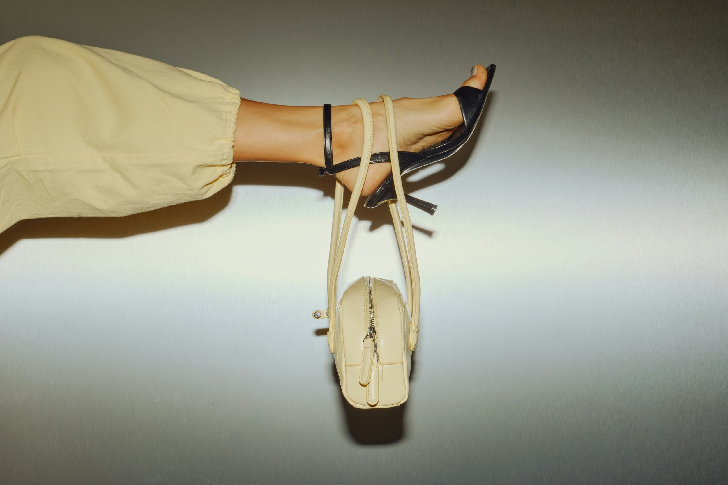 beige pant leg, wearing a black high heel with ankle straps, hanging upside down from a swinging manoeuvre, holding two handbags, one black and one cream-coloured, against a plain wall background. Palette Prescription Colour Analysis