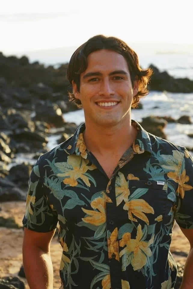 A man smiling on a beach with rocks and water in the background, wearing a colorful floral Hawaiian shirt.