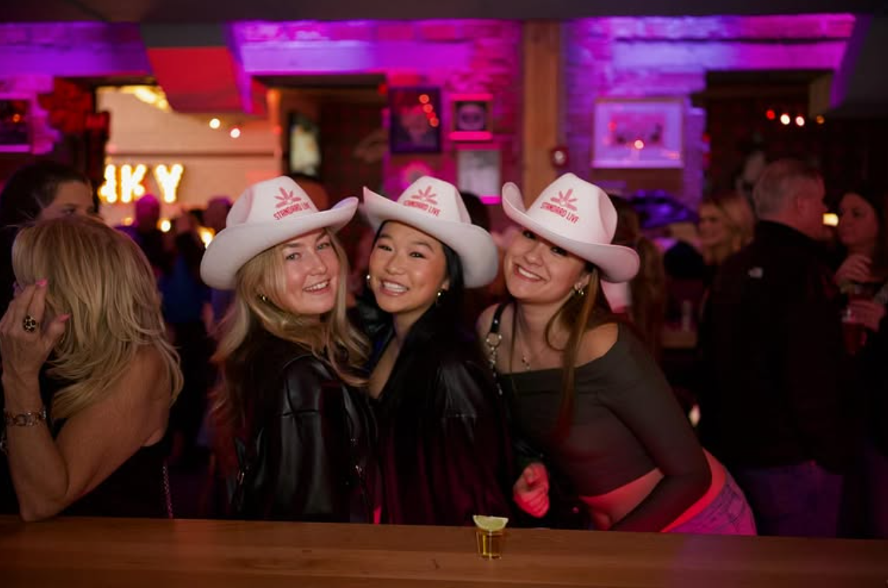 Three women wearing matching white Standard Live branded cowboy hats during a night out.
