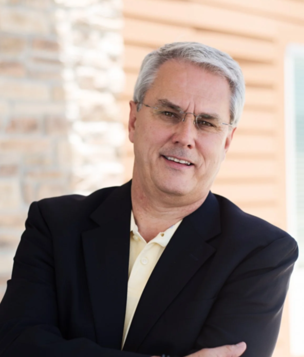 A middle-aged man with gray hair, wearing glasses, a black blazer, and a yellow shirt, smiling with arms crossed in an indoor setting with a brick and wood wall background.