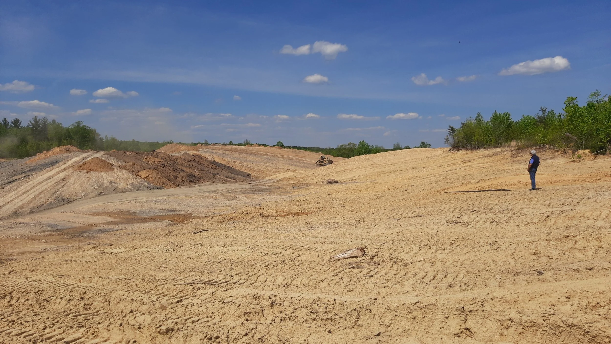 A construction site with dry, cracked earth, a person standing to the right observing, and a small bulldozer in the distance under a blue sky with scattered clouds.