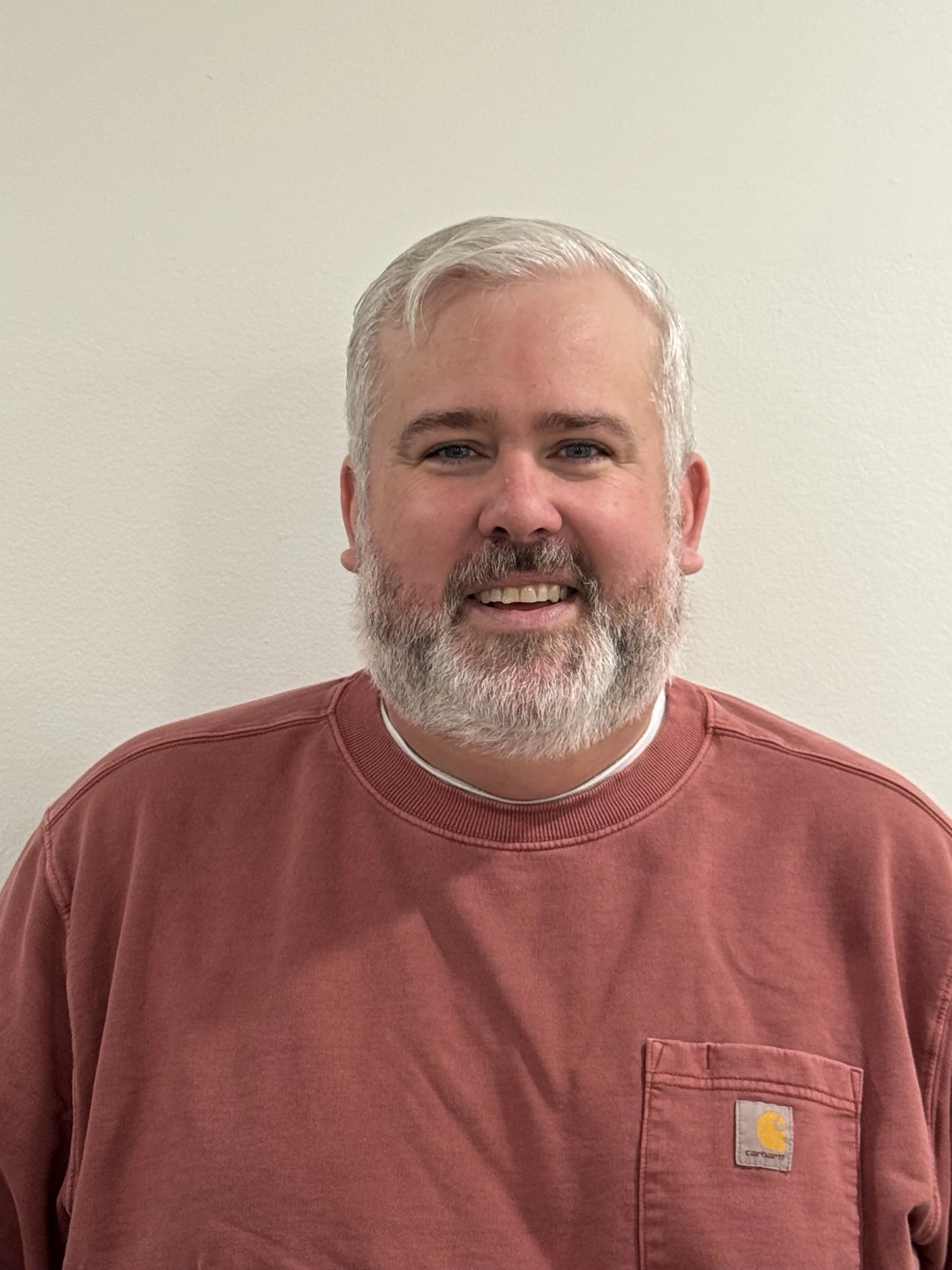 A middle-aged man with a gray beard and hair, smiling, wearing a red Carhartt sweatshirt, standing against a plain light-colored wall.