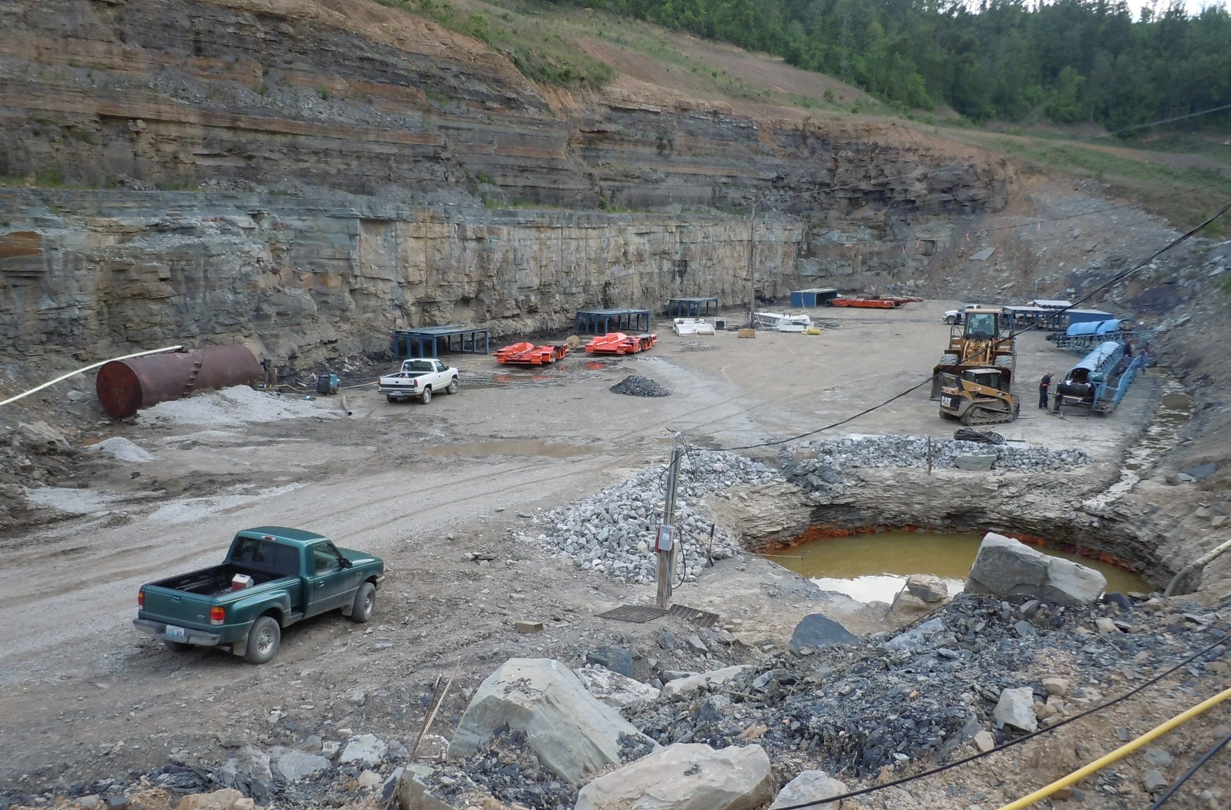 Construction site in a rocky, open area with heavy machinery, trucks, and construction materials, surrounded by dug-out earth and hills.