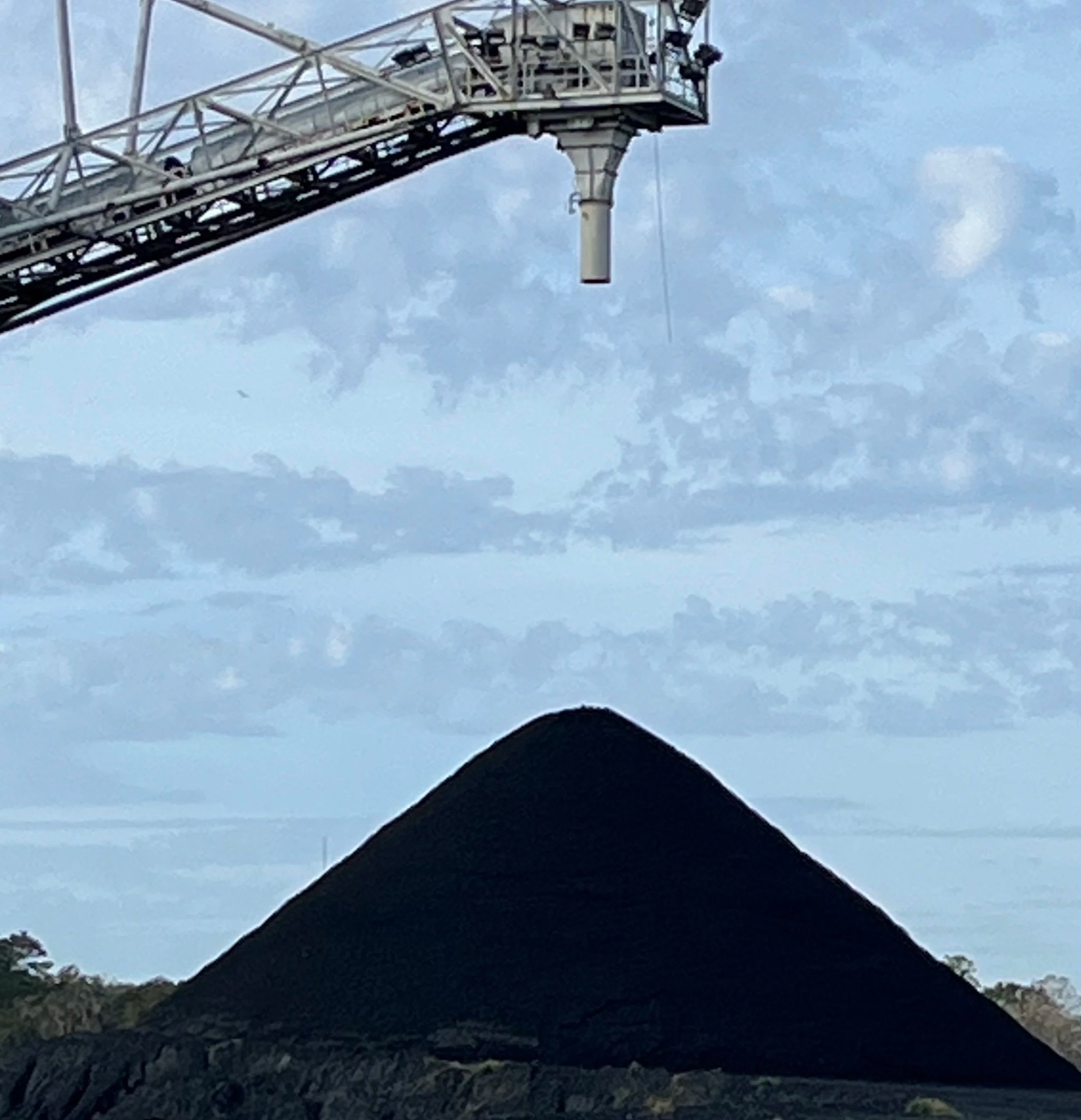 A large, conical mound of dark material with a crane extending above it against a partly cloudy sky.
